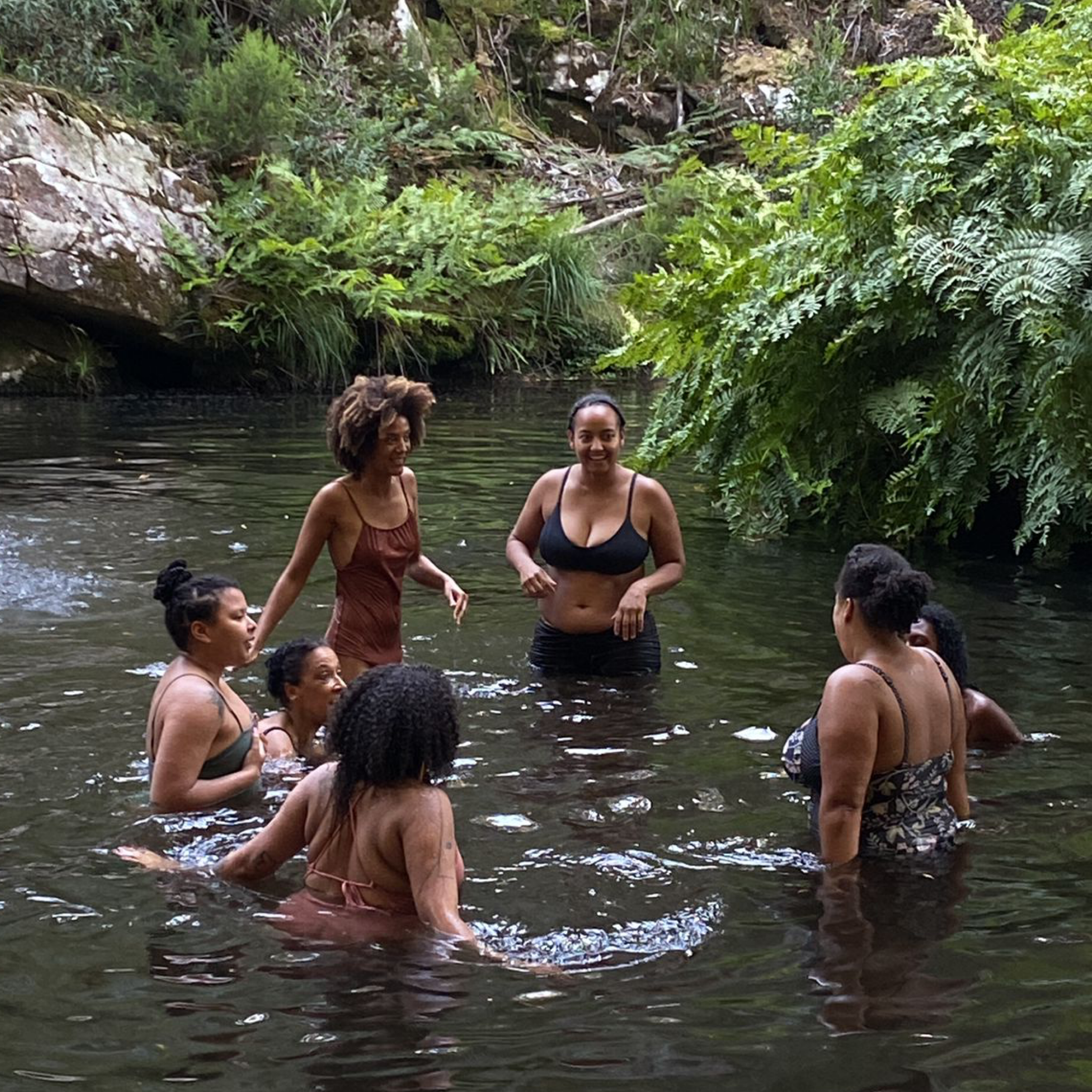 A group of seven women swimming and standing in a natural freshwater stream surrounded by lush green foliage and rocks.