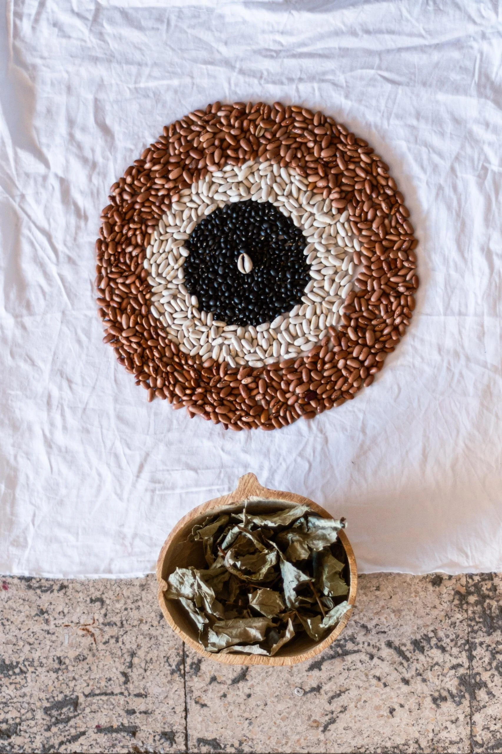 A circular pattern made from different colored beans on a white cloth, with a bowl of dried grape leaves below it on a granite surface.