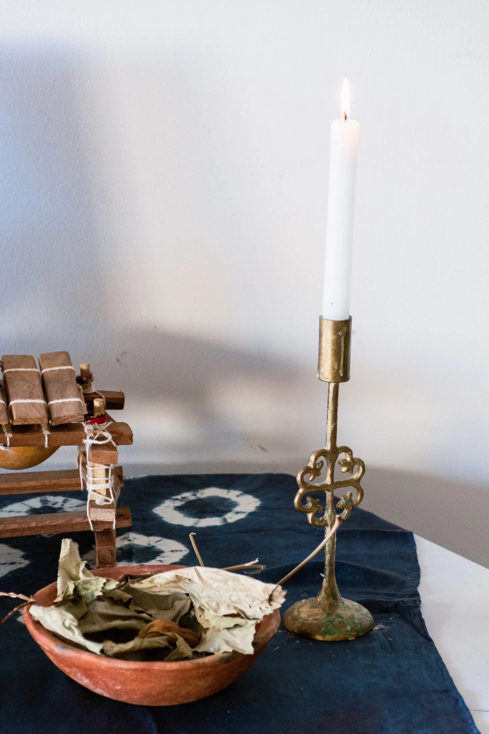 A lit white taper candle in a gold-colored holder, a bowl of dried leaves, and a small wooden boat model on a black cloth, with a plain wall background.