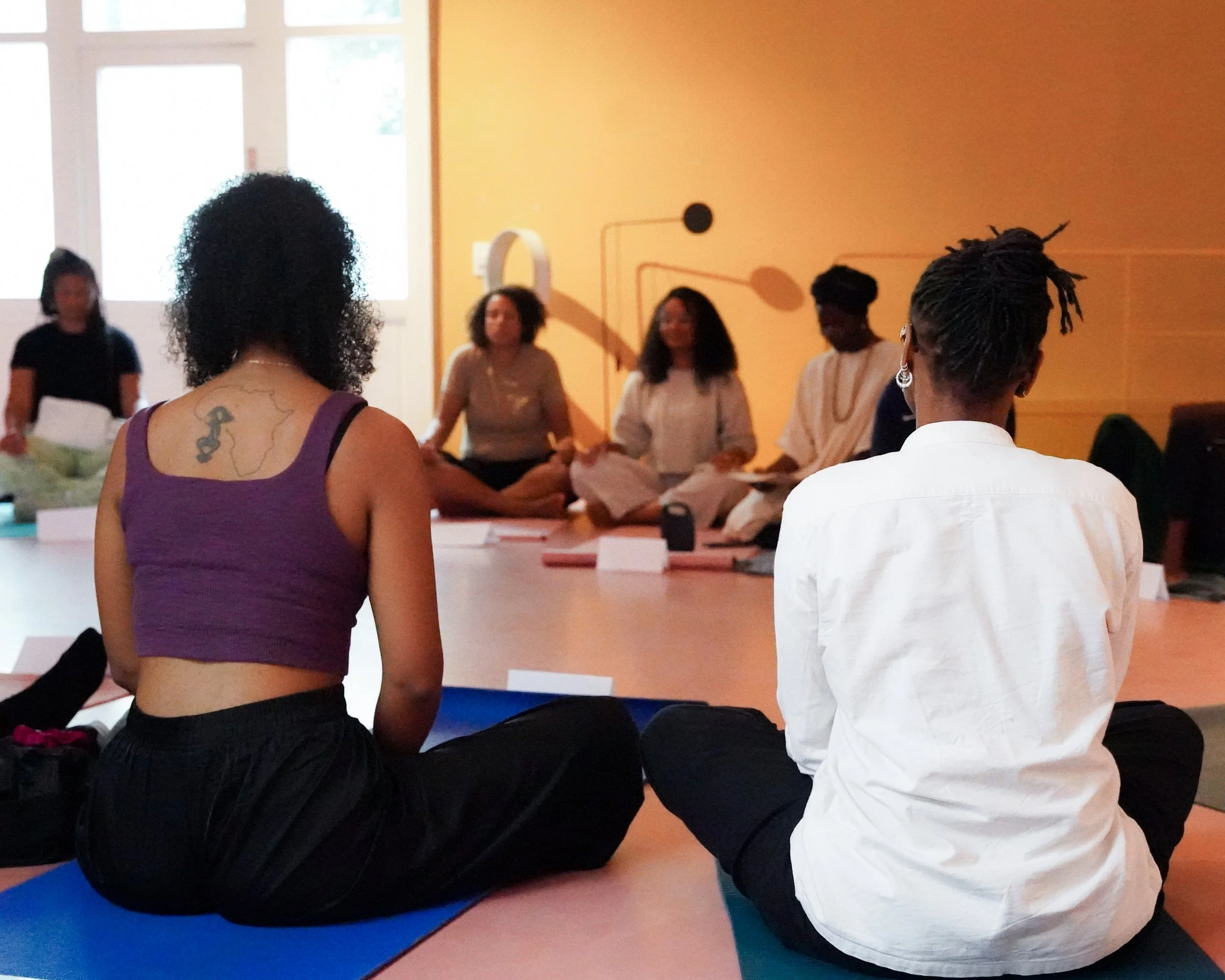 People sitting in a circle on yoga mats during a meditation or yoga class in a room with yellow walls and large windows.