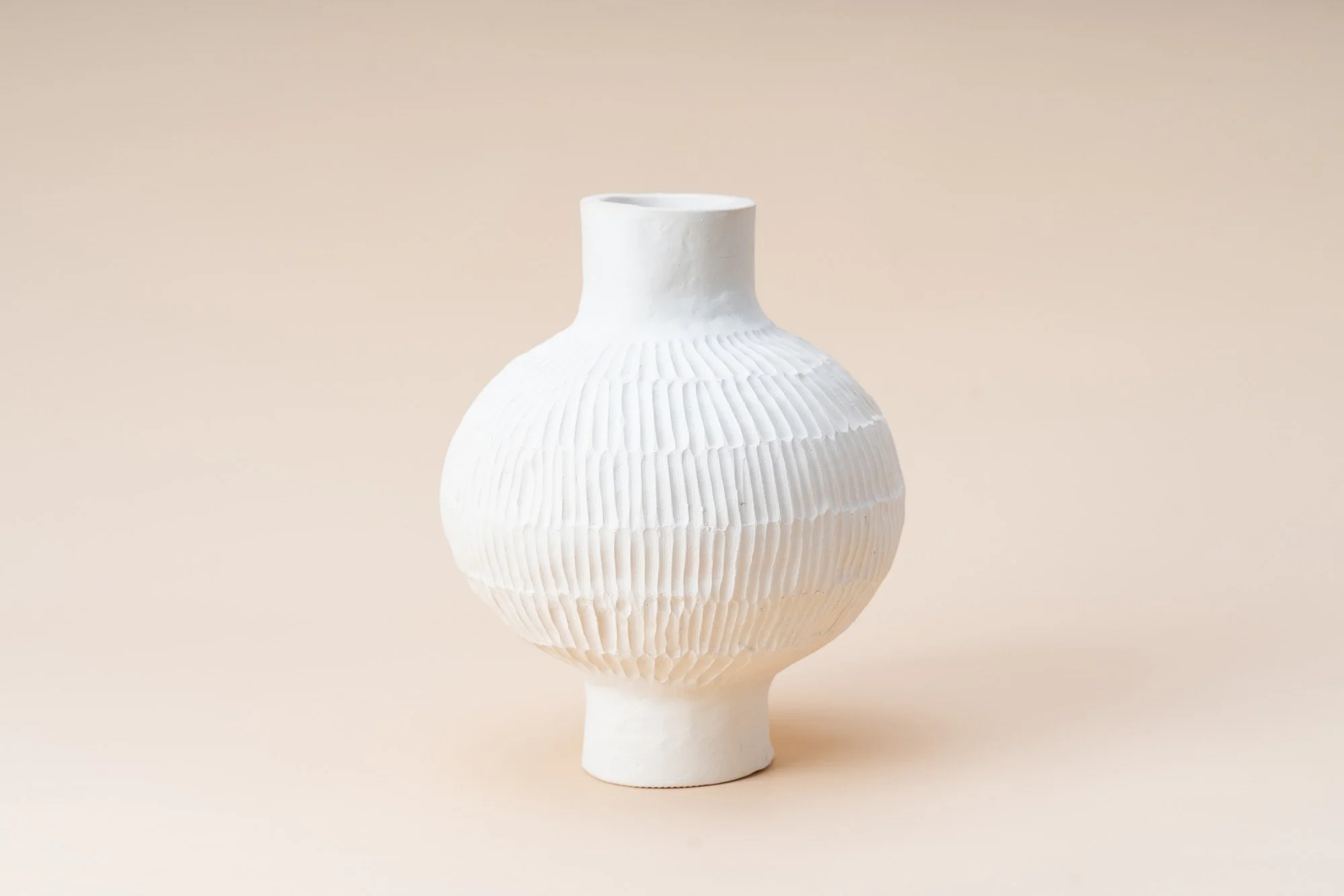 White ceramic vase with textured horizontal ridges, placed on a beige background.