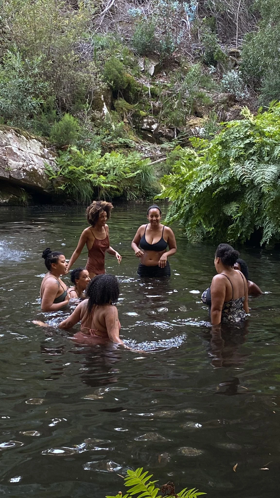 Group of women swimming and chatting in a natural forest creek surrounded by green foliage and rocky banks.