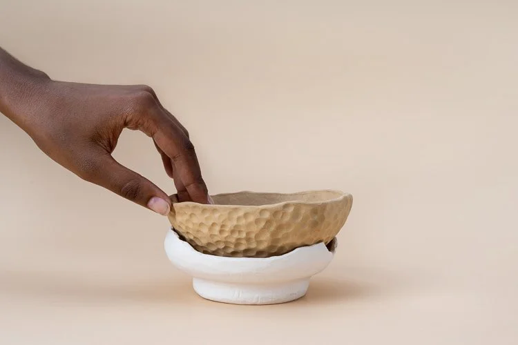 A hand touching a ceramic bowl with a textured, beige interior and a smooth, white exterior, set against a plain beige background.