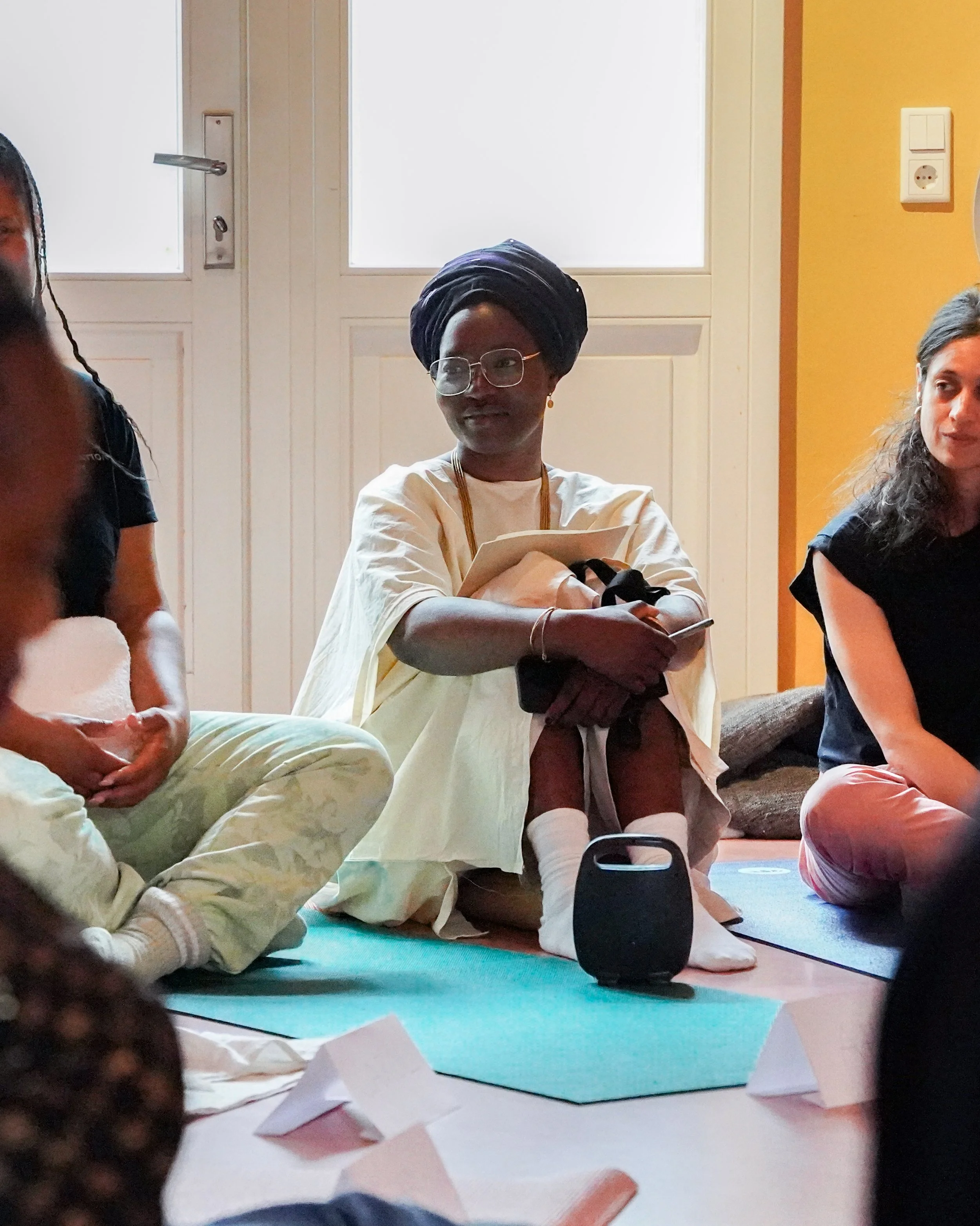 Tobi Aye wearing glasses, a black headwrap, and traditional attire sitting on the floor during a group event.