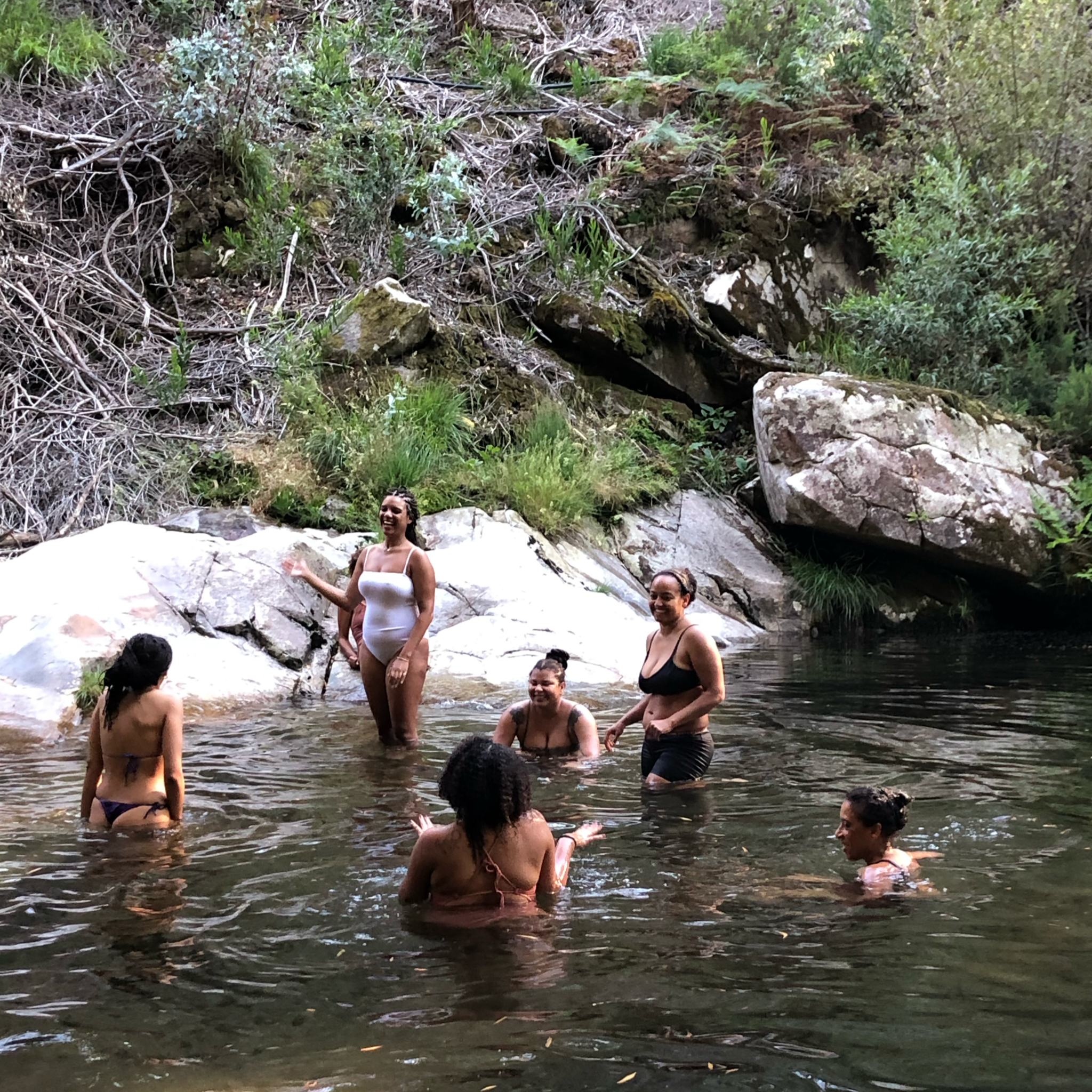 Group of six women enjoying a swim in a natural creek surrounded by rocks and lush green trees.