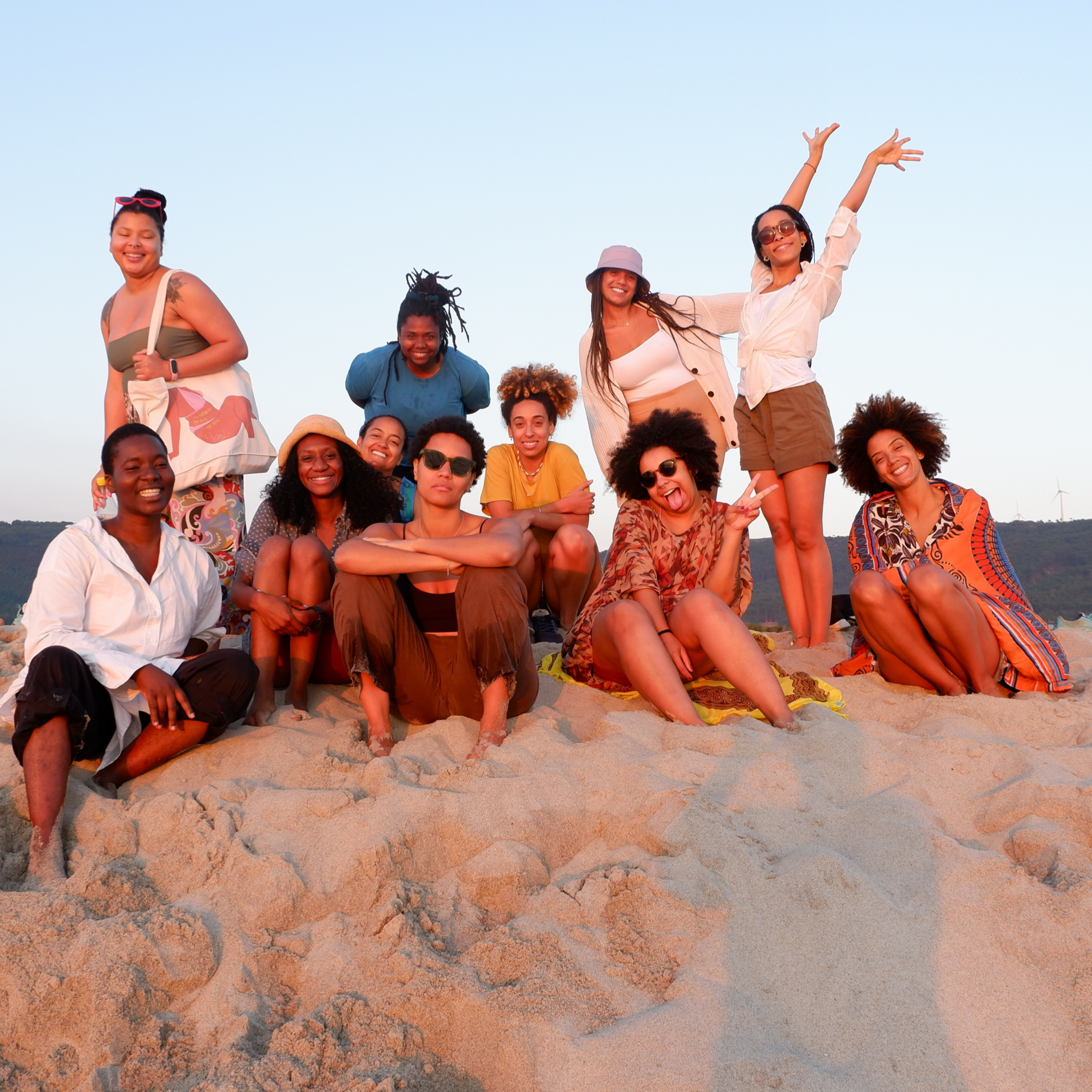 A group of twelve women enjoying a sunny day at the beach, sitting and standing on sand, with a clear sky and distant hills in the background.