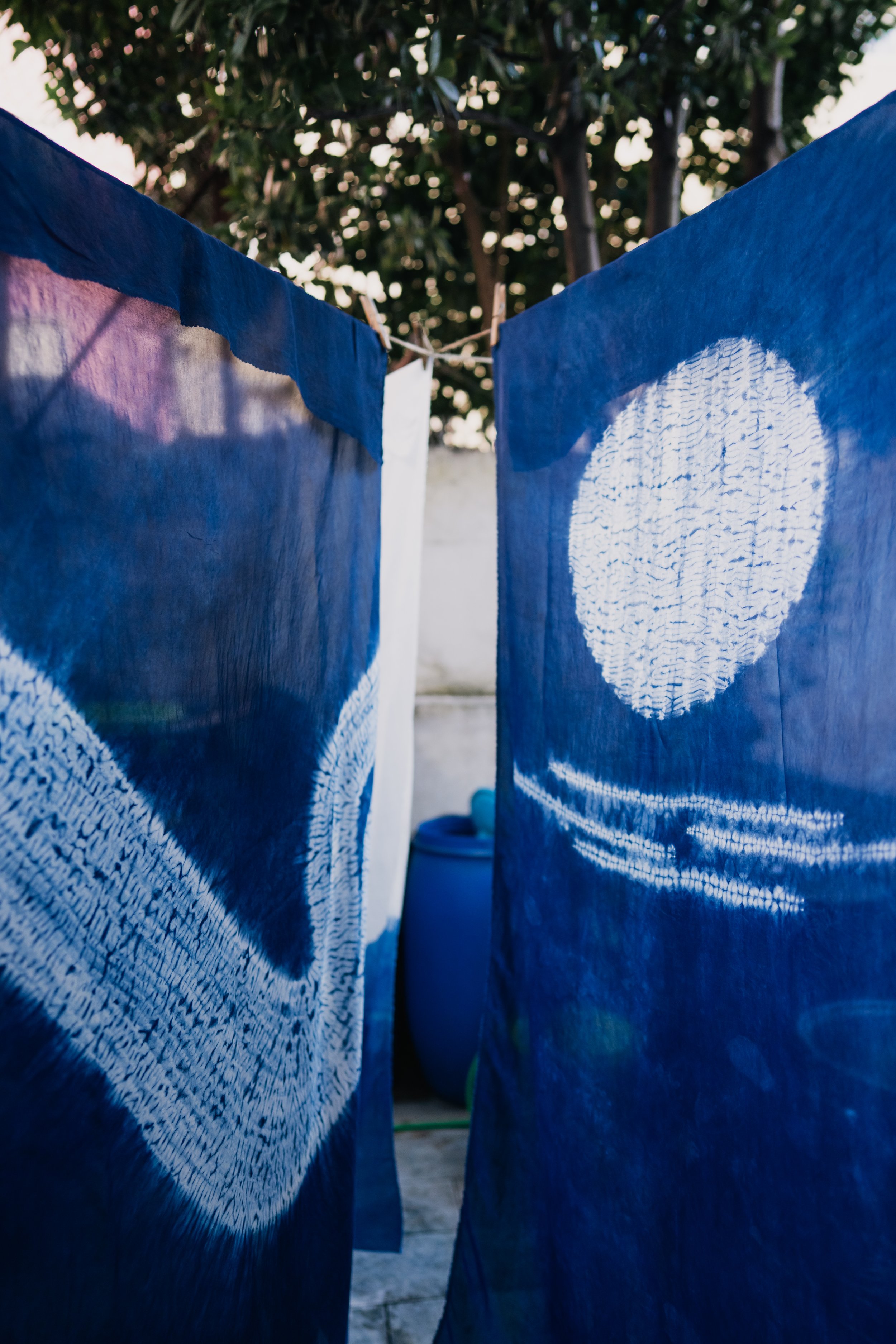 Blue tie-dye fabric hanging on a clothesline with white painted patterns of a circle and wavy lines, outdoors with trees and a blue barrel in the background.