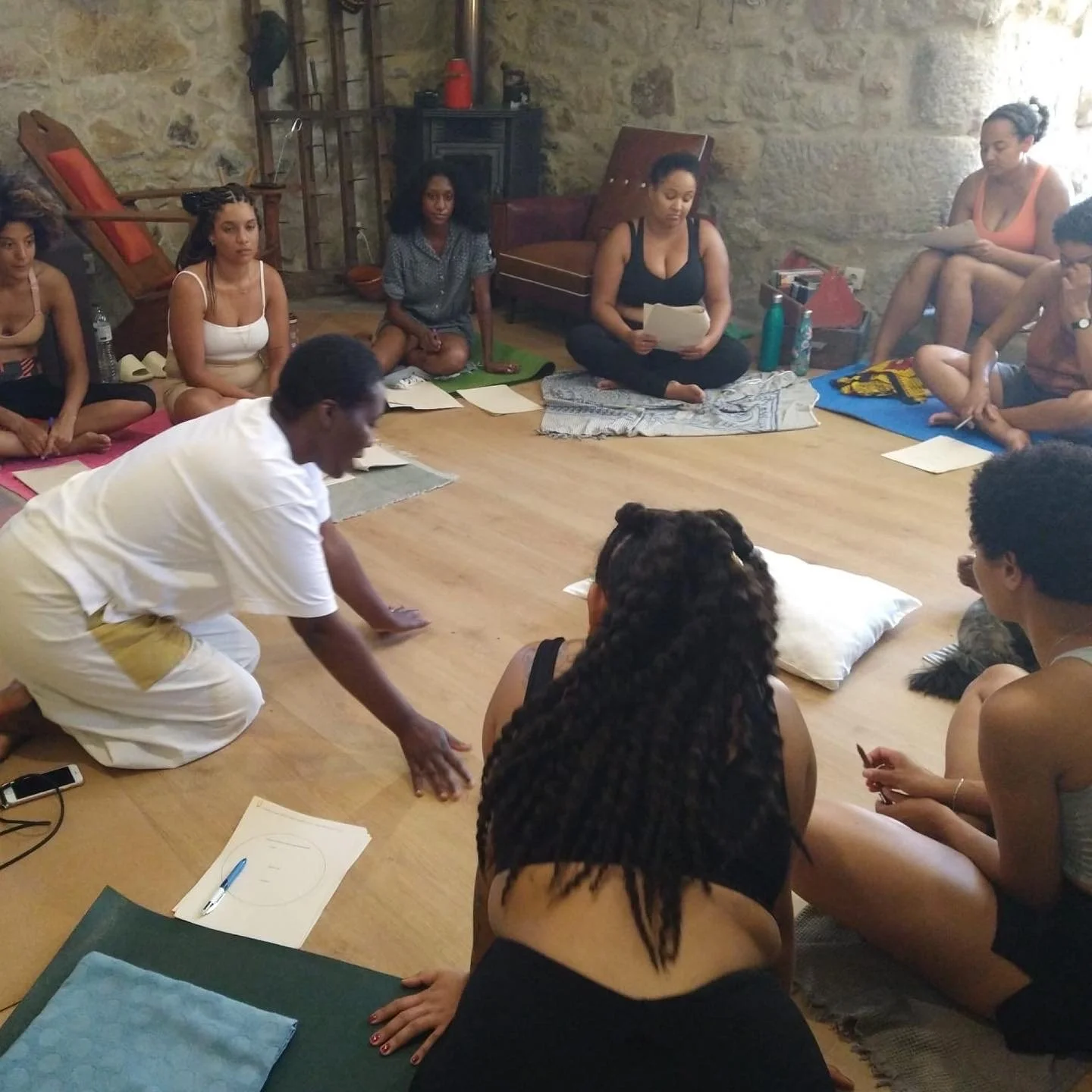 Group of people sitting on the floor in a circle, participating in a workshop or class with papers, notebooks, and a man leading the activity.