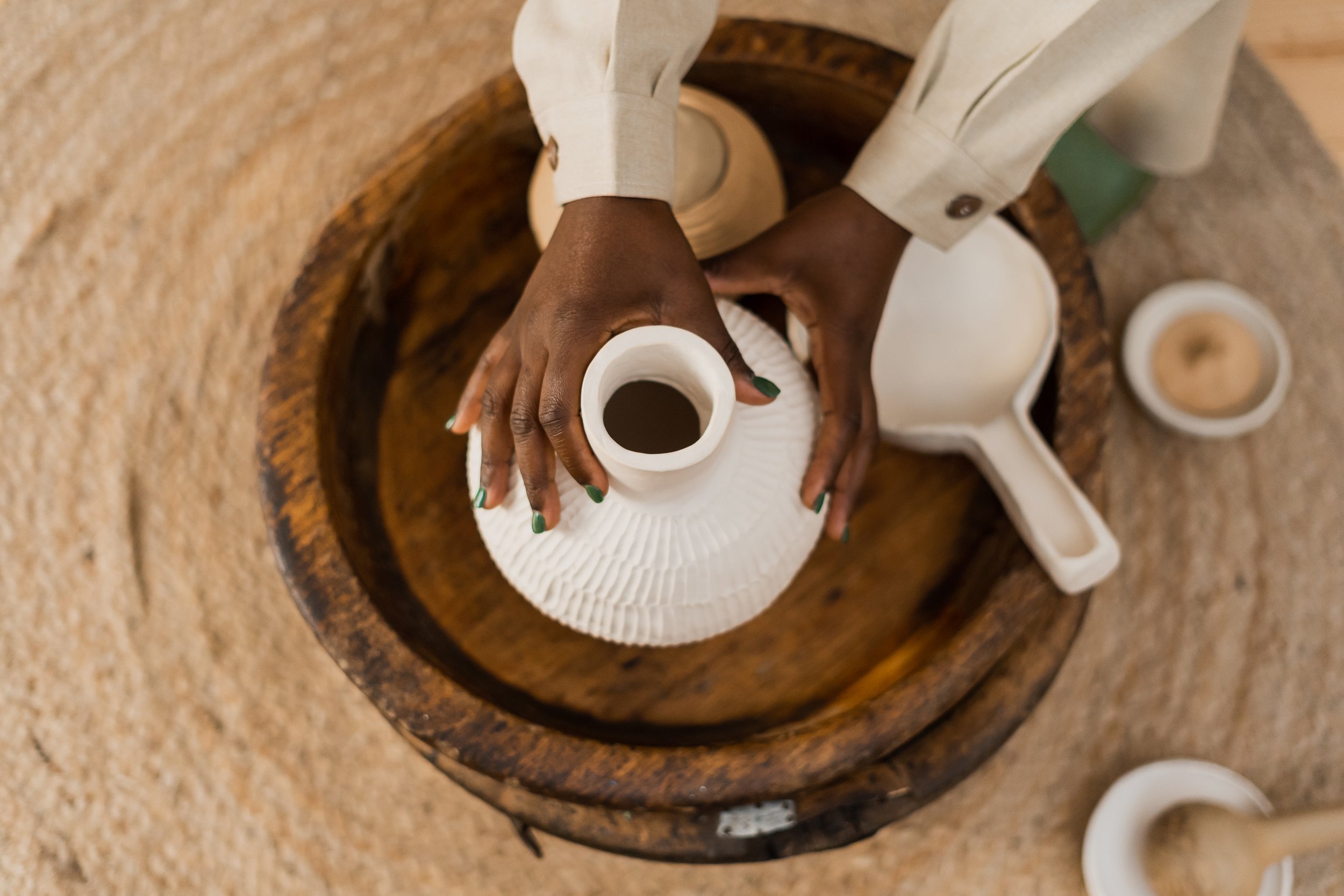 Person arranging white ceramic vessels inside a wooden bowl on a sandy surface.