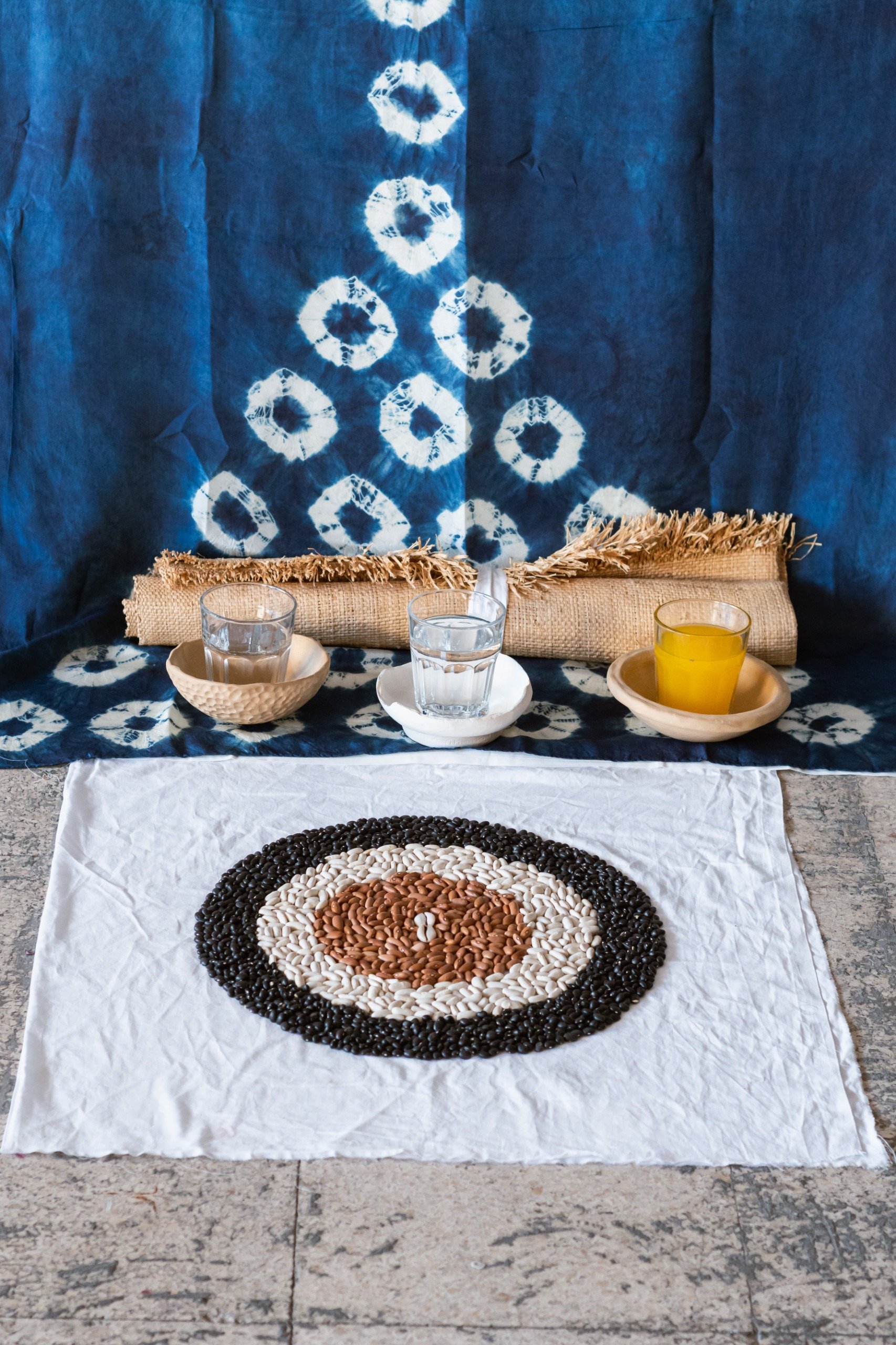A display of black, white, and brown beans arranged in a circular pattern on a white cloth. Behind the beans, there is a blue fabric backdrop with white tie-dye circles. In front of the backdrop, there are three glasses of water and one glass of yell