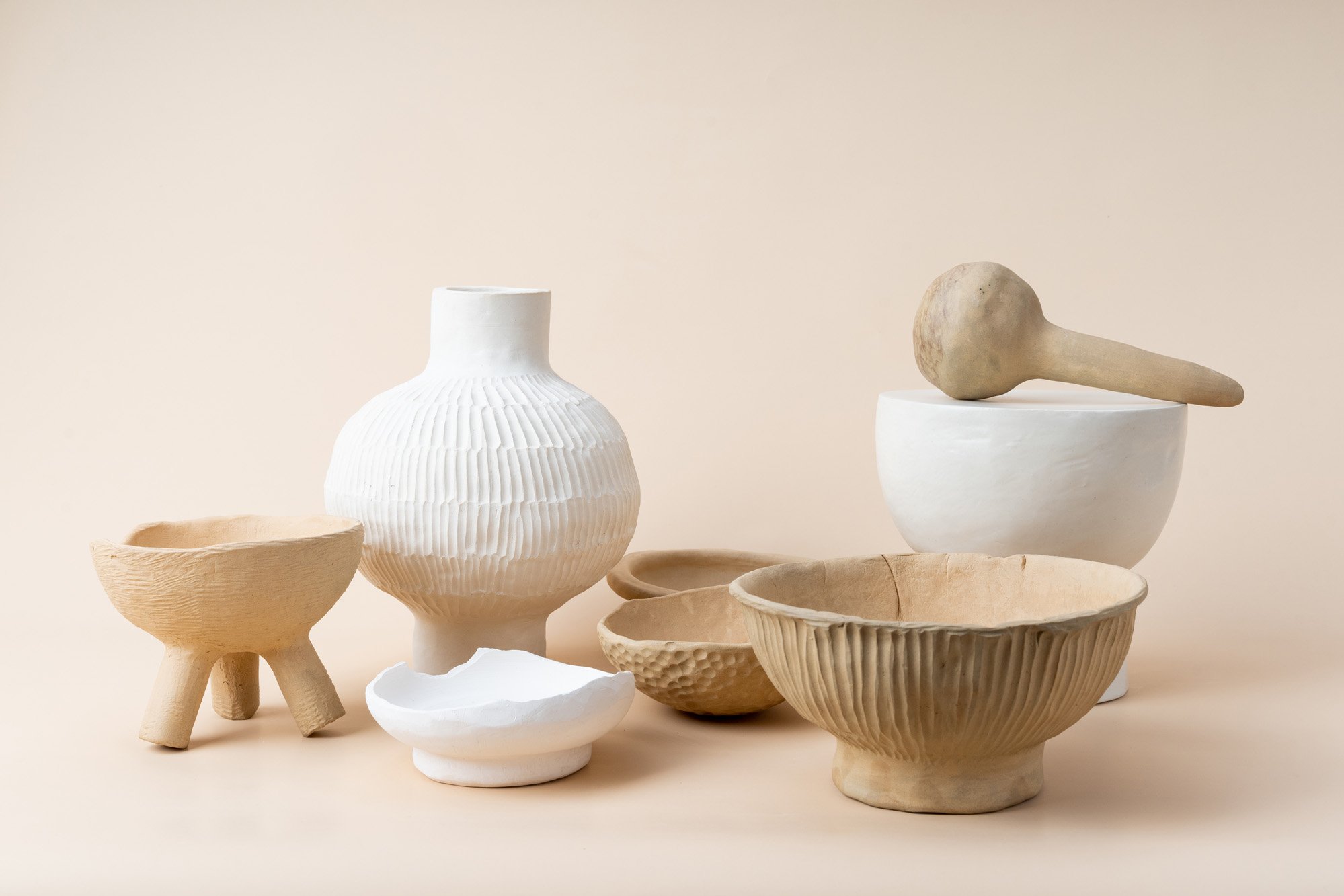 Various beige and white ceramic and clay bowls and vases on a neutral background, including a bowl with three legs, a tall textured vase, and a mortar and pestle.