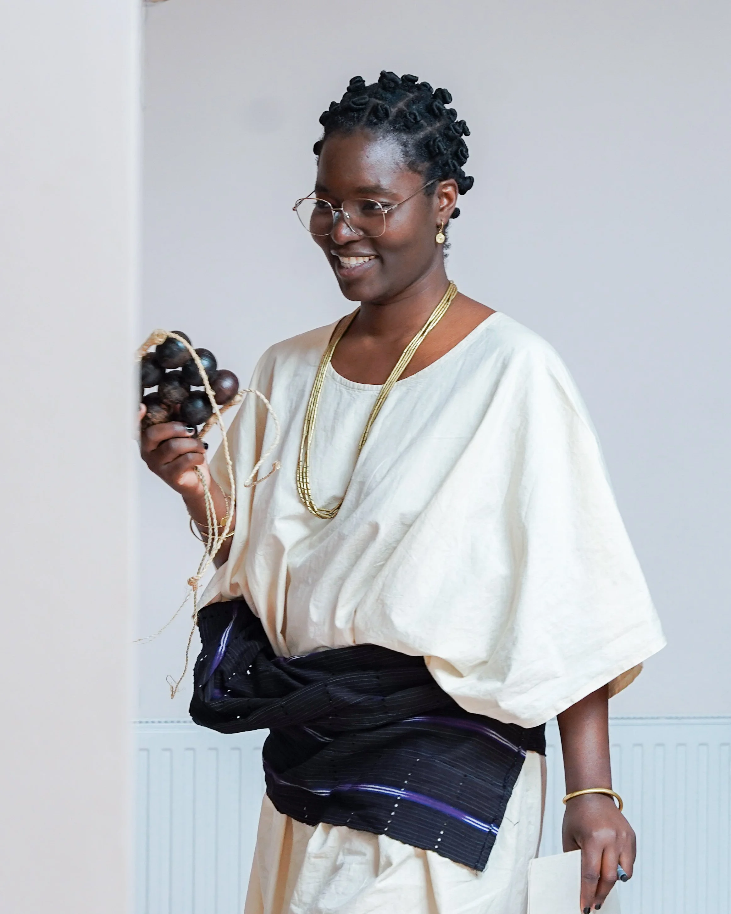 A smiling woman with glasses and short, coiled hair, wearing a cream-colored dress, gold jewelry, and a black patterned sash, holding a string of black beads in her right hand, standing indoors against a plain wall.