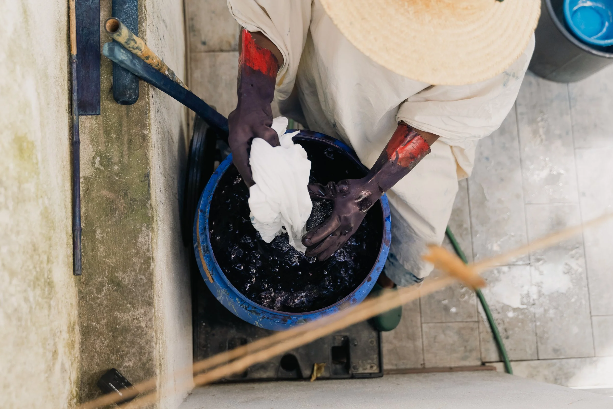 A person wearing gloves and a wide-brimmed hat is washing clothes in a blue bucket.