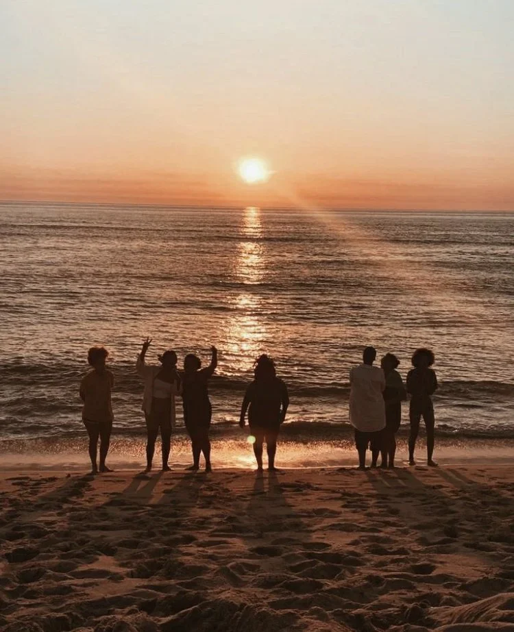 Silhouettes of eight people standing on a beach during sunset, with the ocean and horizon in the background.