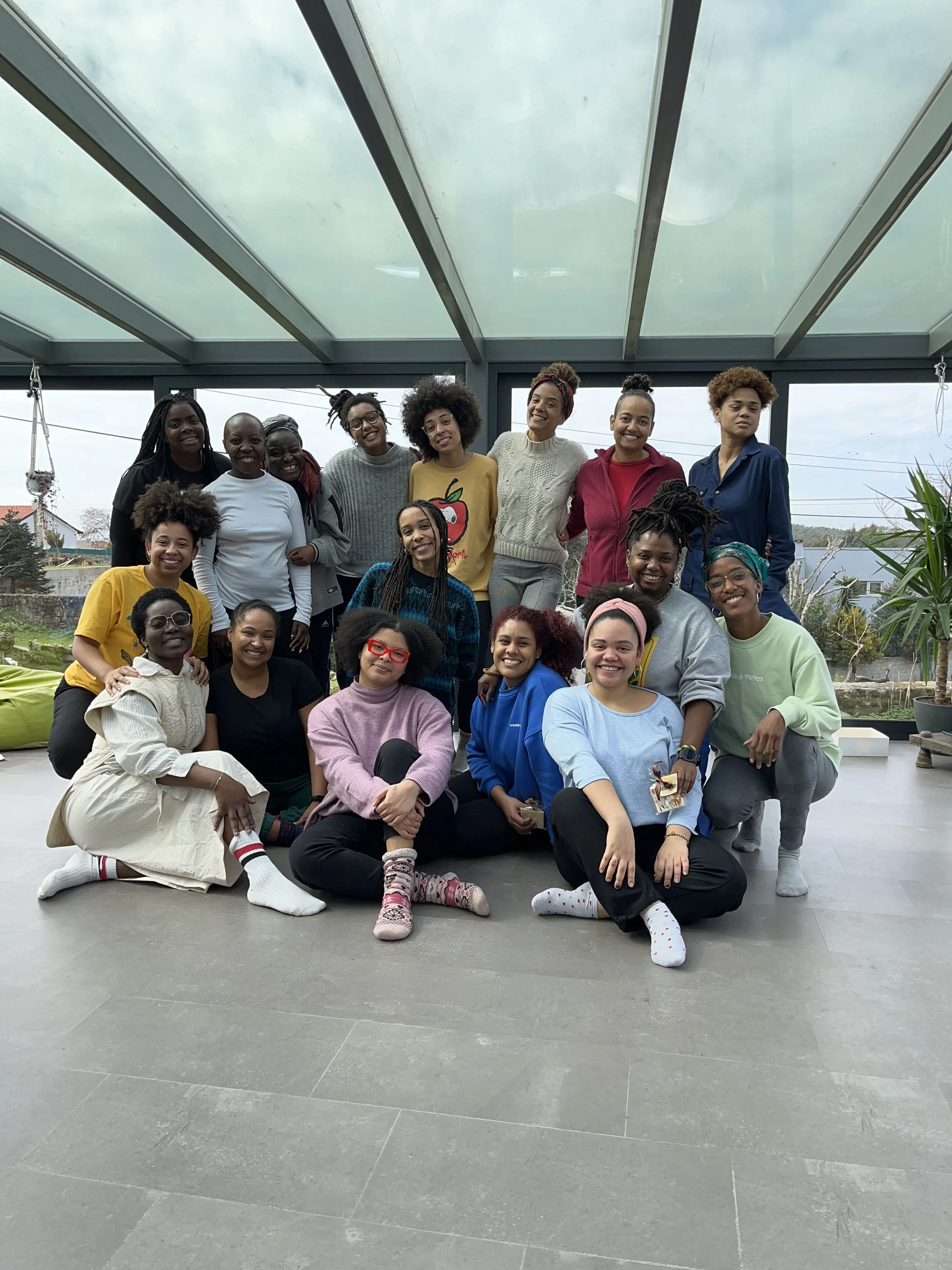 A group of people posing together on a large, bright porch with a glass ceiling, outdoor scenery, and potted plants in the background.