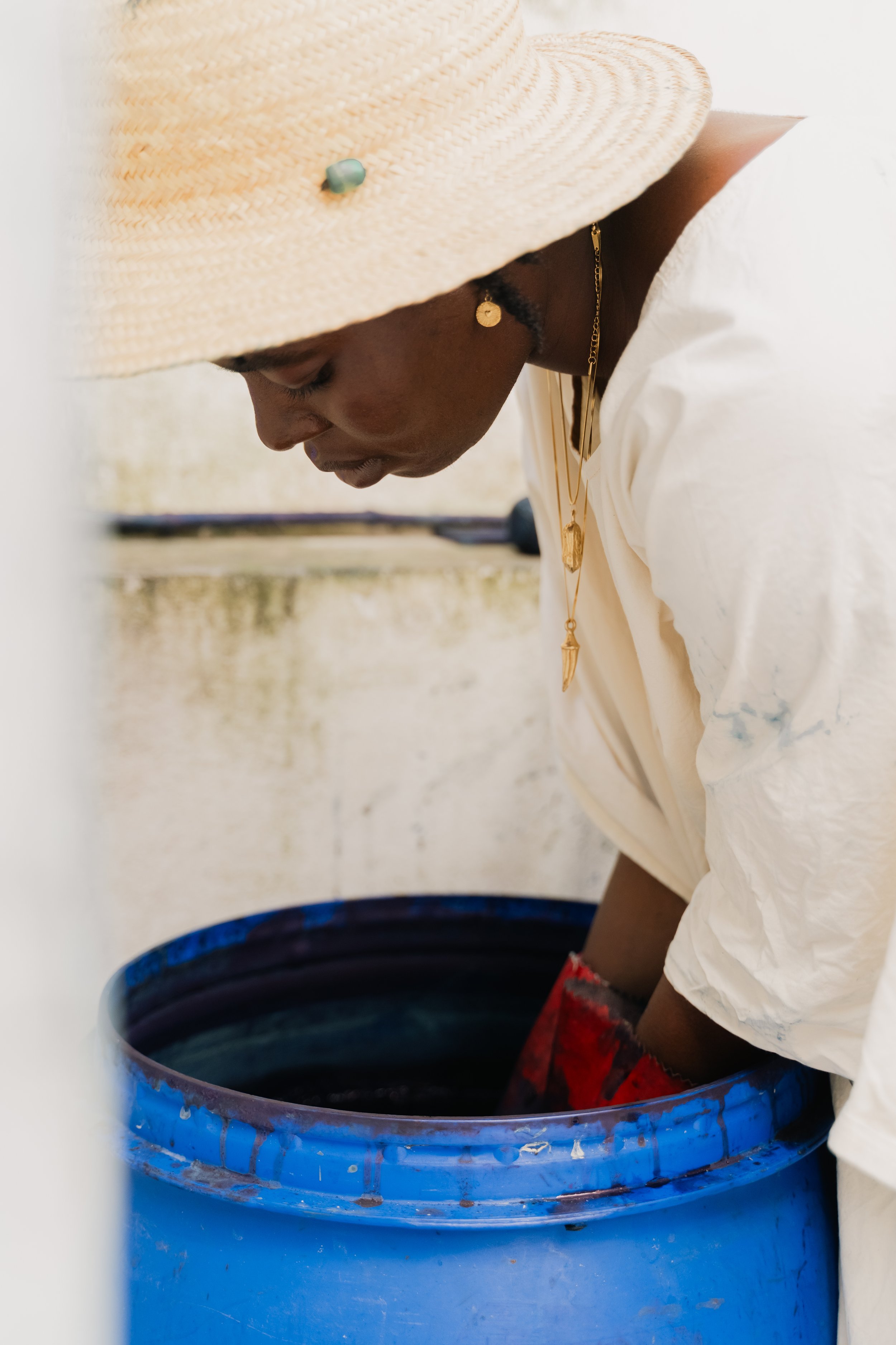 A person wearing a large straw hat, gold jewelry, and a white shirt dipping their hand into a large blue barrel filled with dark liquid.