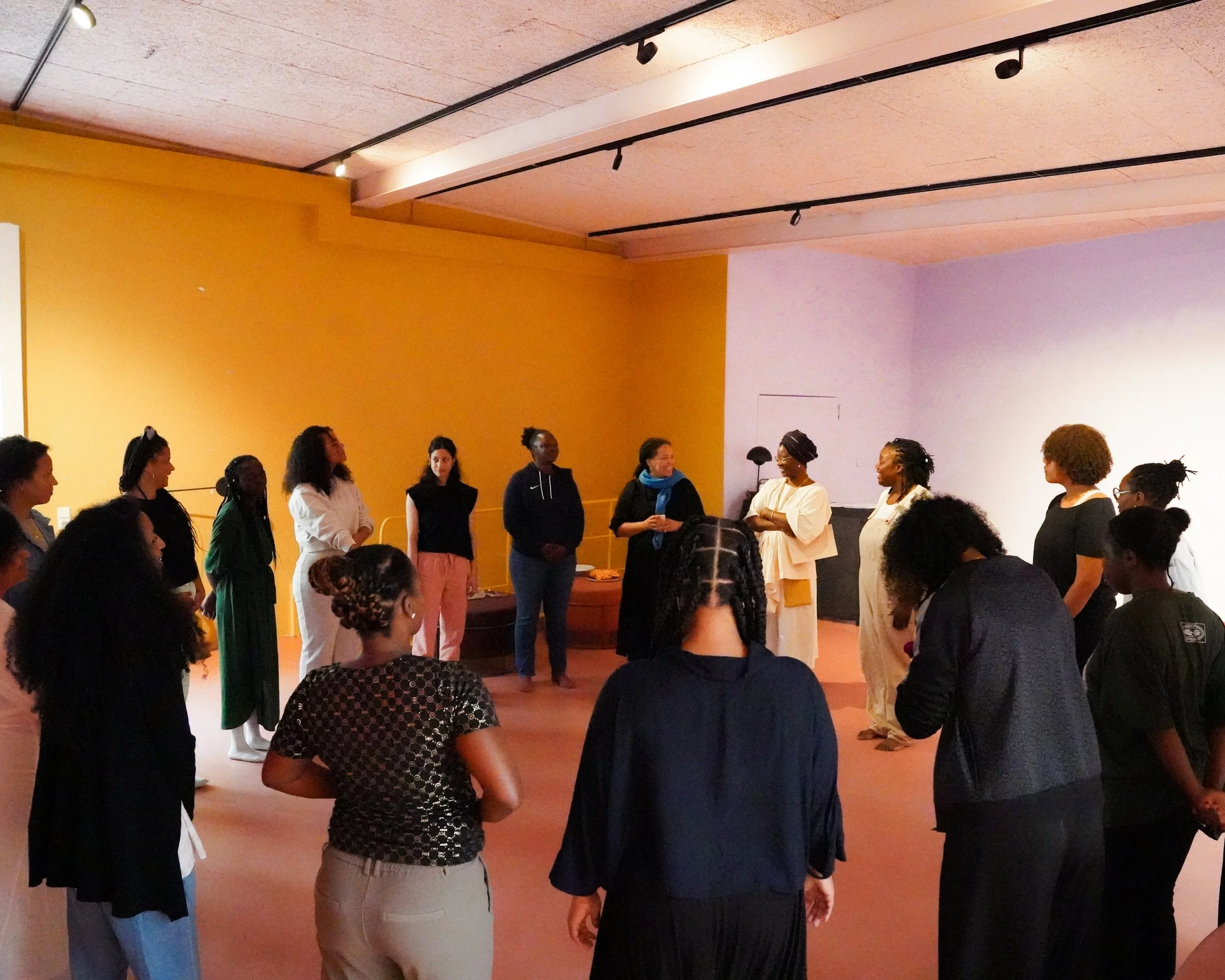 A group of women standing in a circle indoors, engaged in conversation or activity, in a room with yellow and purple walls and wooden ceiling.