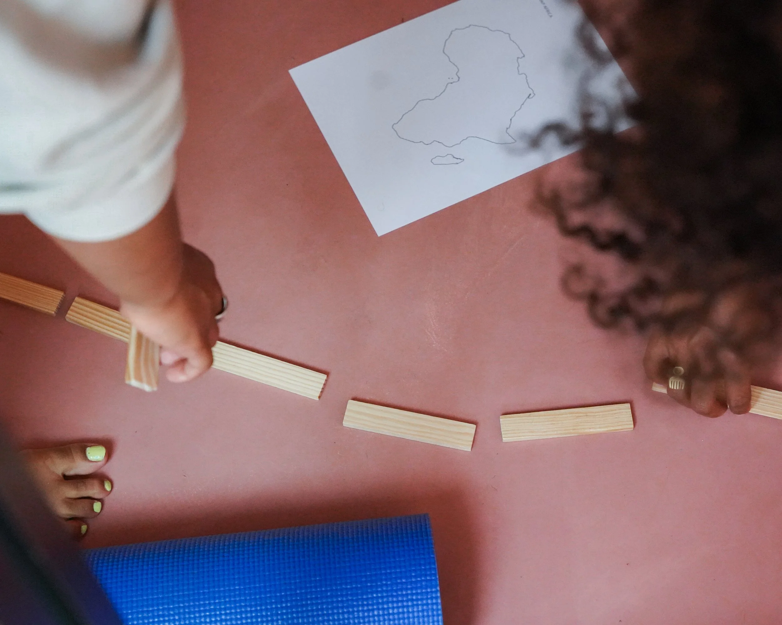 Two people are arranging horizontal wooden planks on a reddish-pink surface, with a blue yoga mat and a white paper with an outline of a country or island in the background. One person's hand is visible with painted nails, and the other person's curl