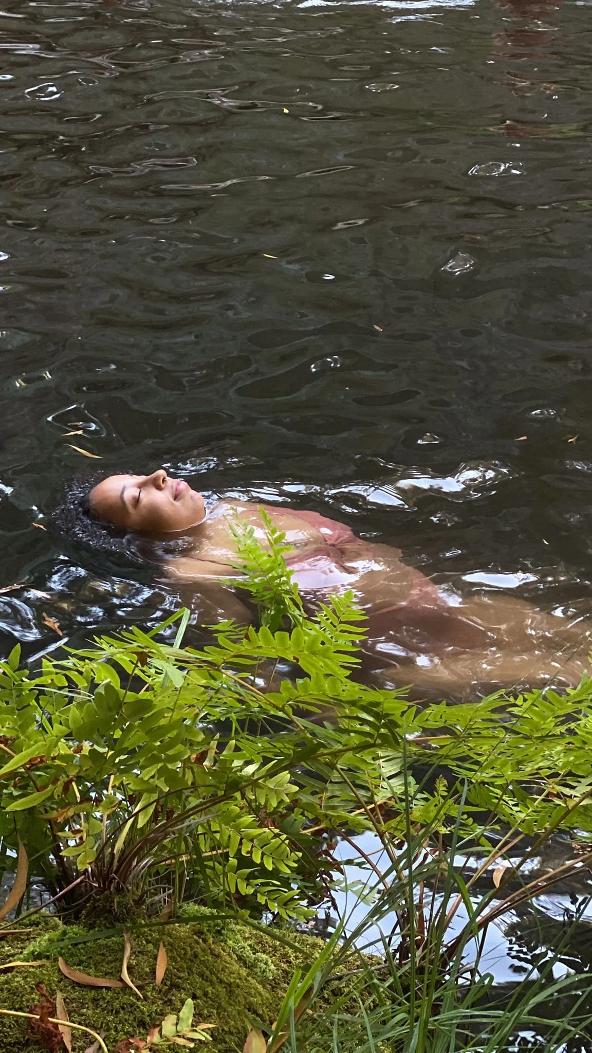 A woman with eyes closed floating peacefully in a river surrounded by green plants.