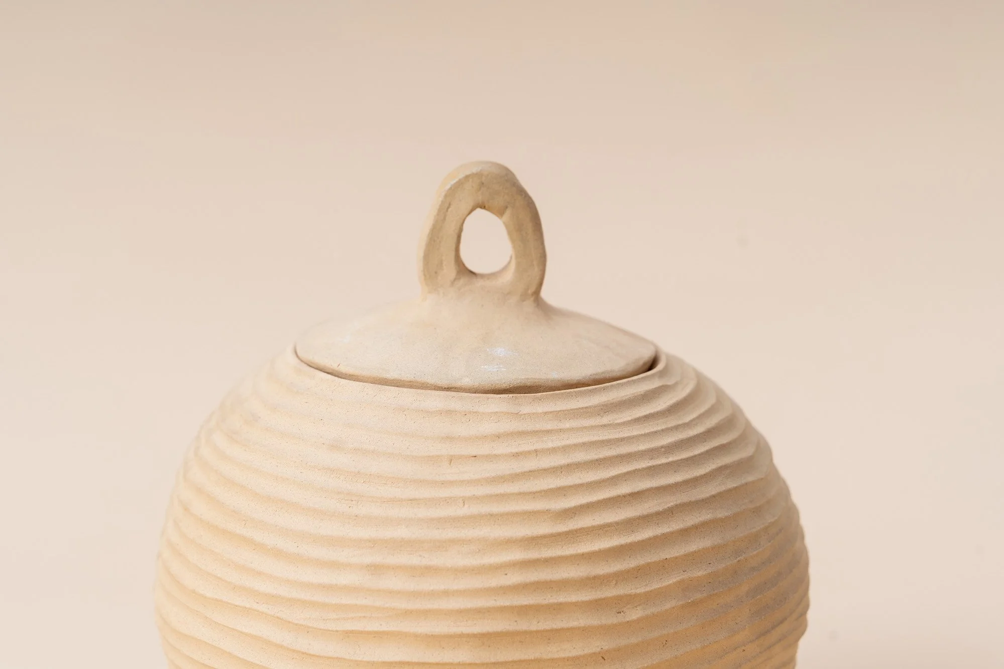 Close-up of a beige ceramic jar with concentric ridges and a small loop handle on the lid.