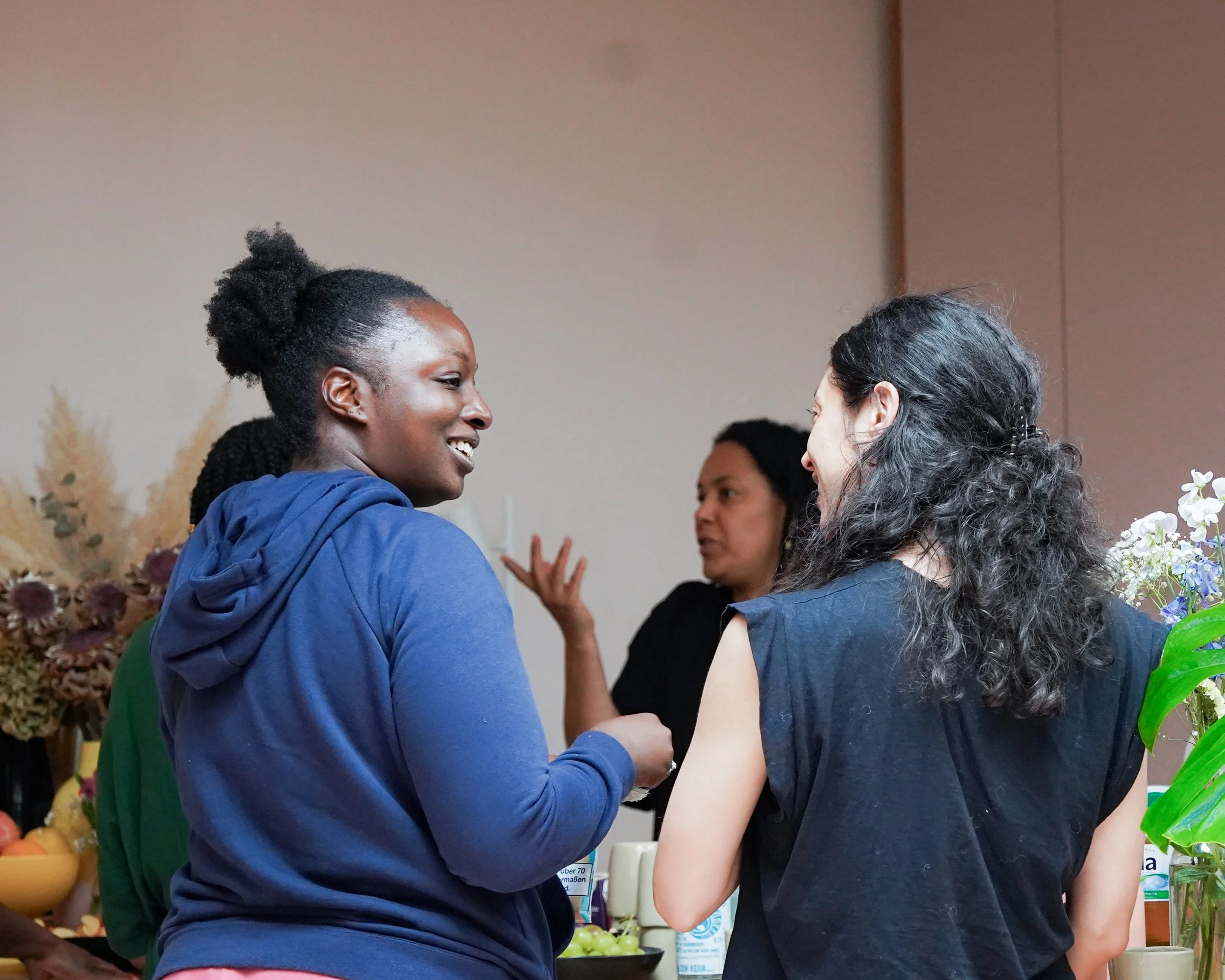 Three women are engaged in conversation at a social gathering, with one woman smiling and wearing a blue hoodie, another woman with dark curly hair, and a third woman in black in the background, with flowers and refreshments on a table behind them.