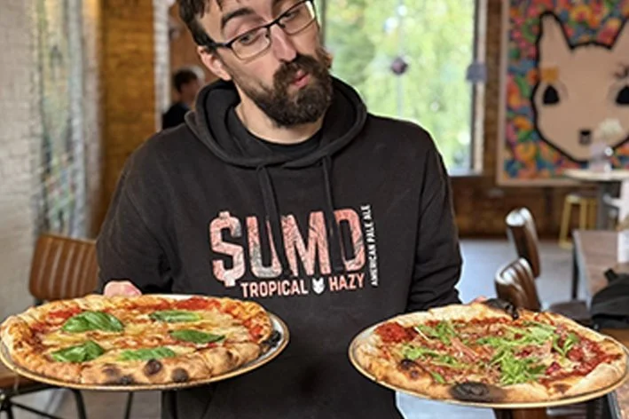Andy wearing a black hoodie holding two pizzas with basil and green toppings
