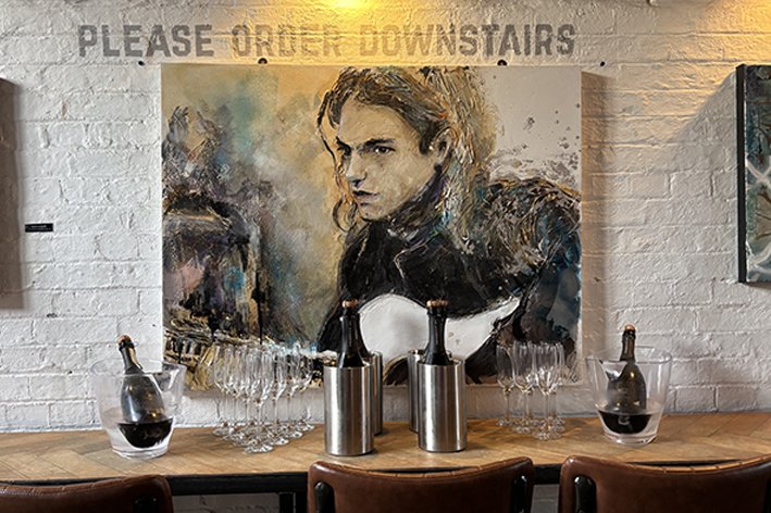 A large painting of a woman with long, wavy hair and intense expression hangs on a white brick wall, above a wooden table with wine bottles, glasses, and ice buckets, and a sign that reads 'Please Order Downstairs'.