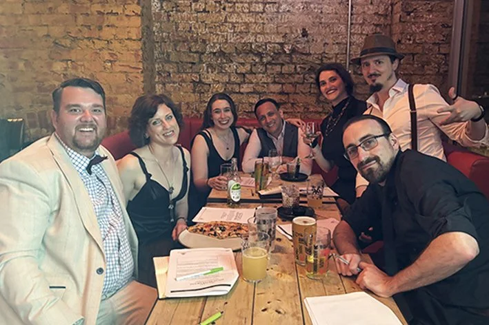 Group of seven people gathered around a wooden table, smiling and enjoying drinks and pizza.
