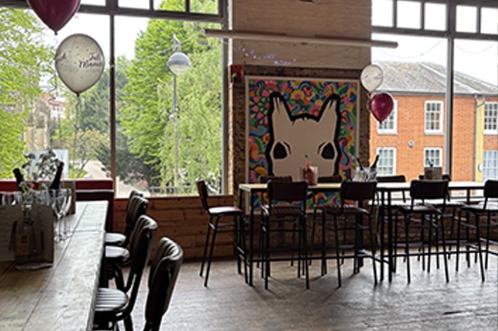Interior of the upstairs room with wooden tables and chairs, balloons, and large windows showing greenery and brick buildings outside.
