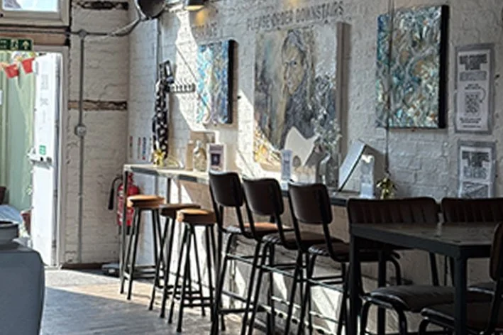 Interior of a café or art studio with a row of black barstools along a white counter, artwork on the brick wall, and a black table with chairs in the foreground.