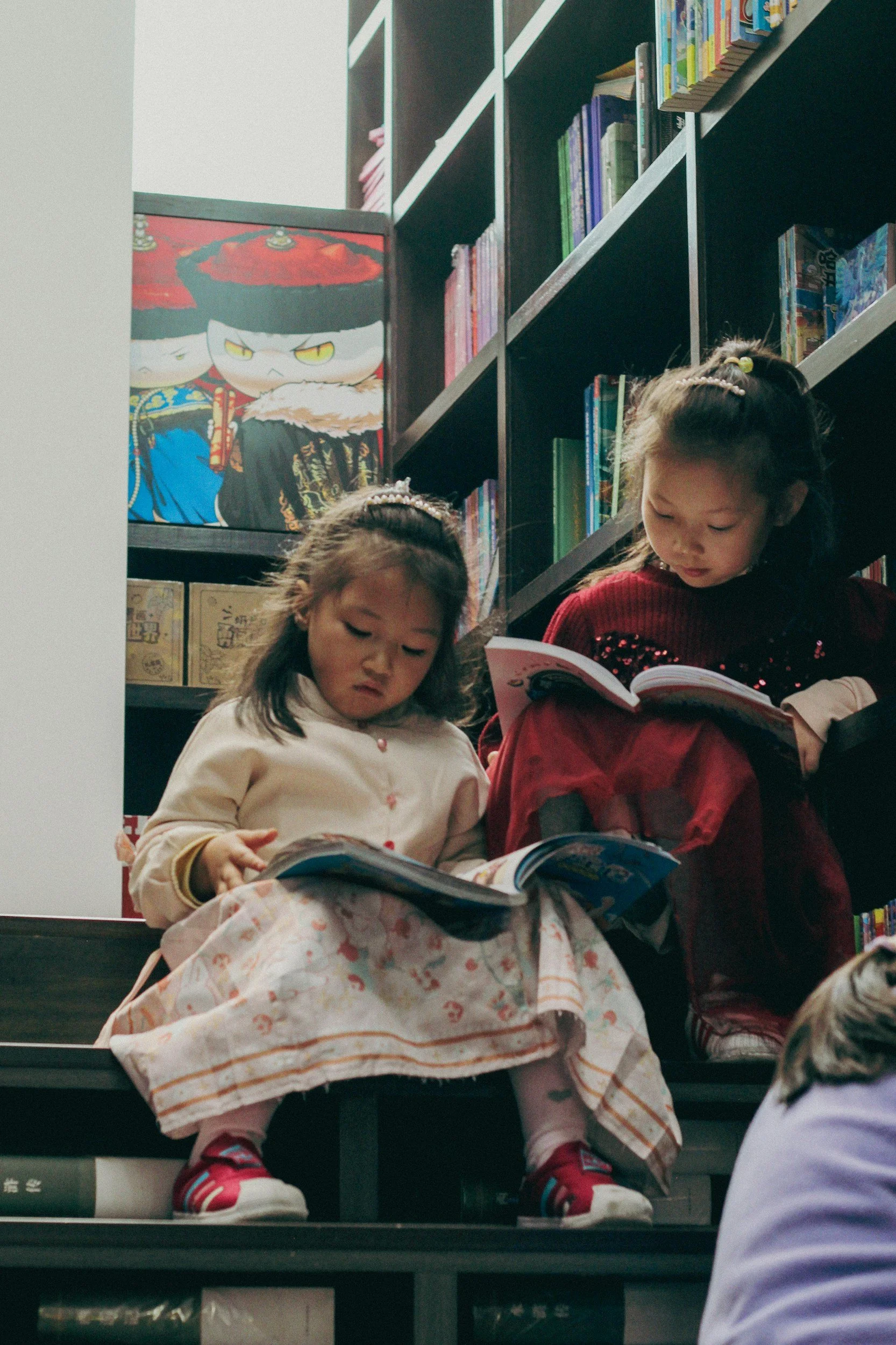 Two young girls sat on the steps in a library with books on their laps, reading