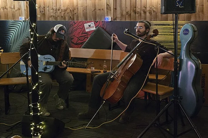 Two musicians playing string instruments, one on guitar and the other on cello, in a cozy indoor setting with wooden walls and decorative lighting.