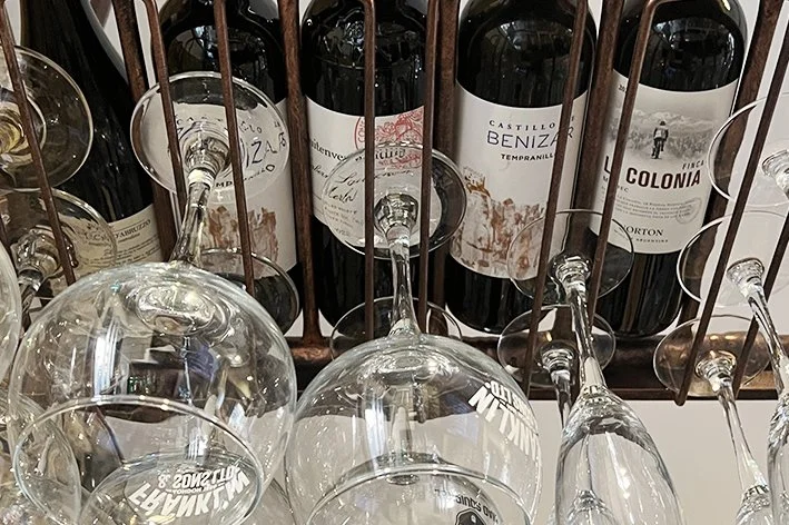 Multiple wine bottles stored behind a vertical metal wine rack with empty wine glasses hanging upside down in front.