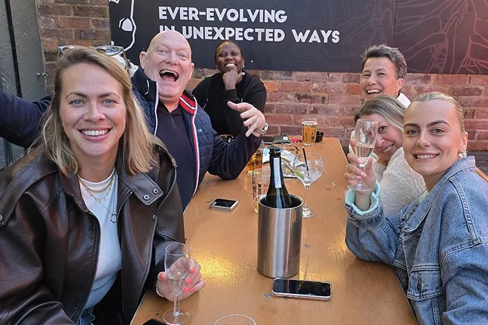 Group of six friends sitting around a table at a bar, smiling and celebrating with drinks and champagne, in a lively setting with a brick wall and a sign in the background.