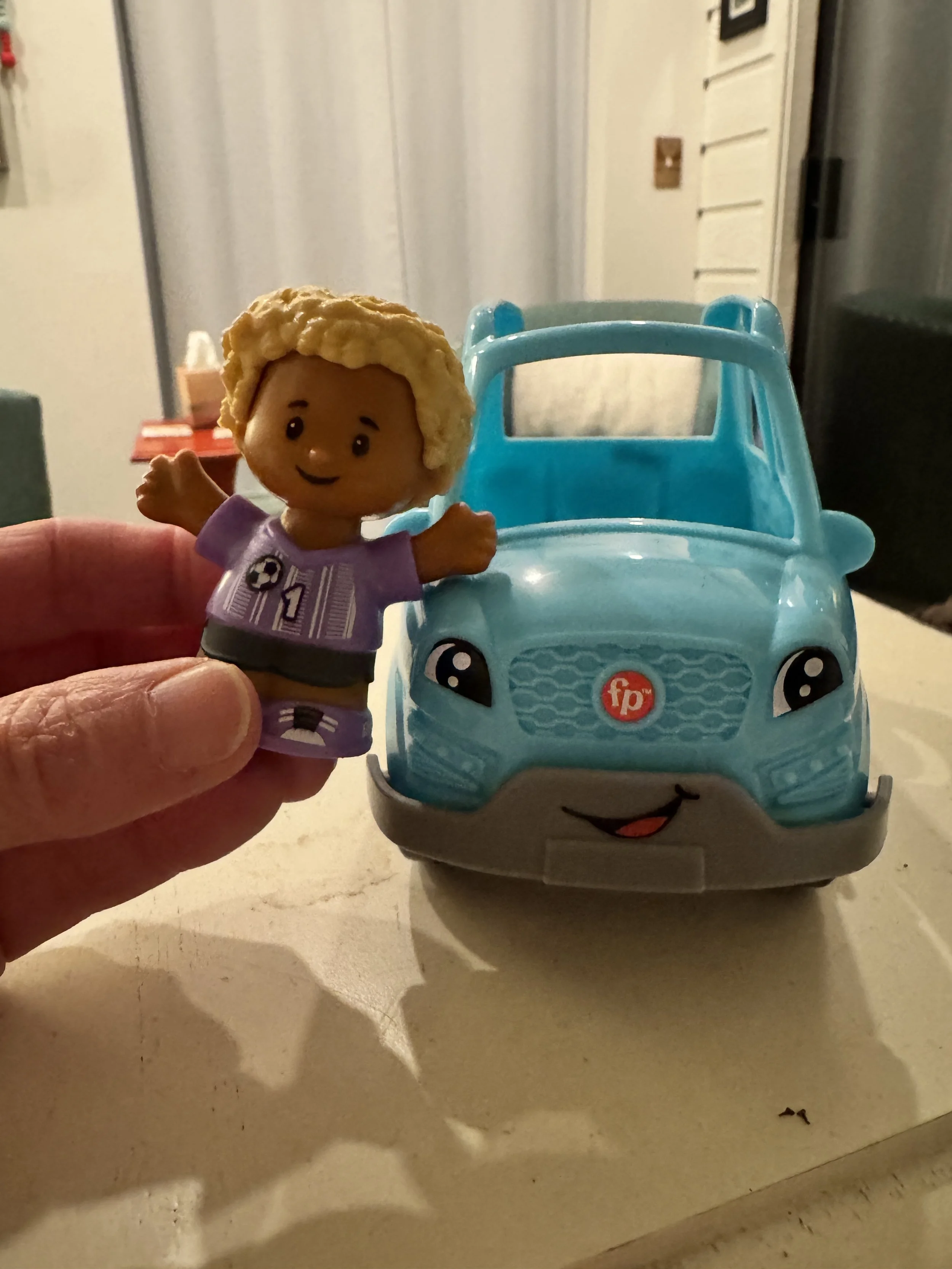 Fisher Price toy boy in soccer shirt standing beside a blue toy car.