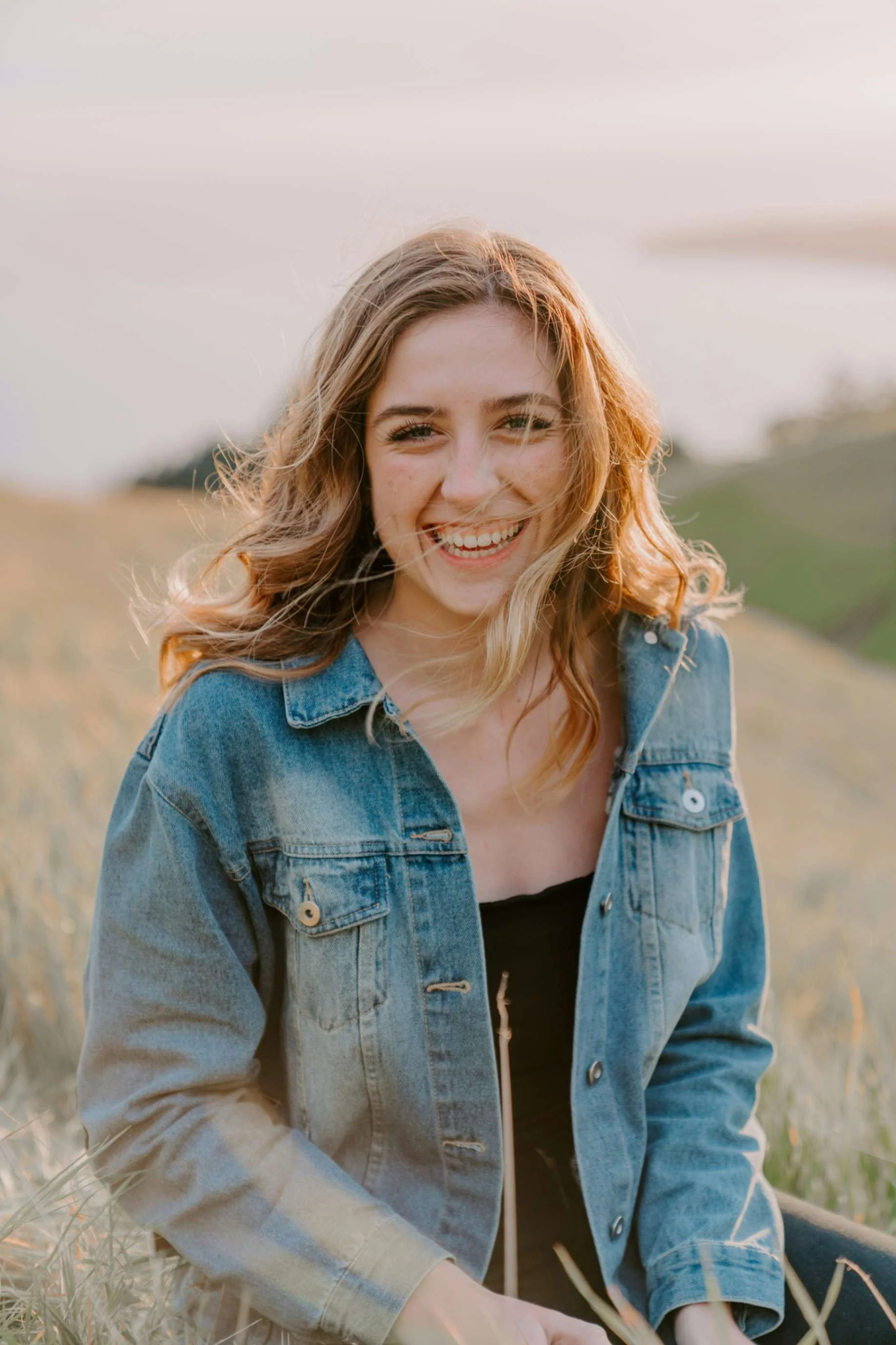 A young woman with wavy, reddish-blonde hair smiling outdoors, wearing a denim jacket and black top, in a grassy field during late afternoon or early evening.