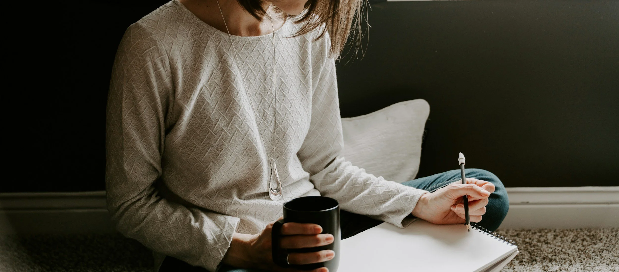 woman sitting on floor writing in her journal with a cup of coffee in her hand.