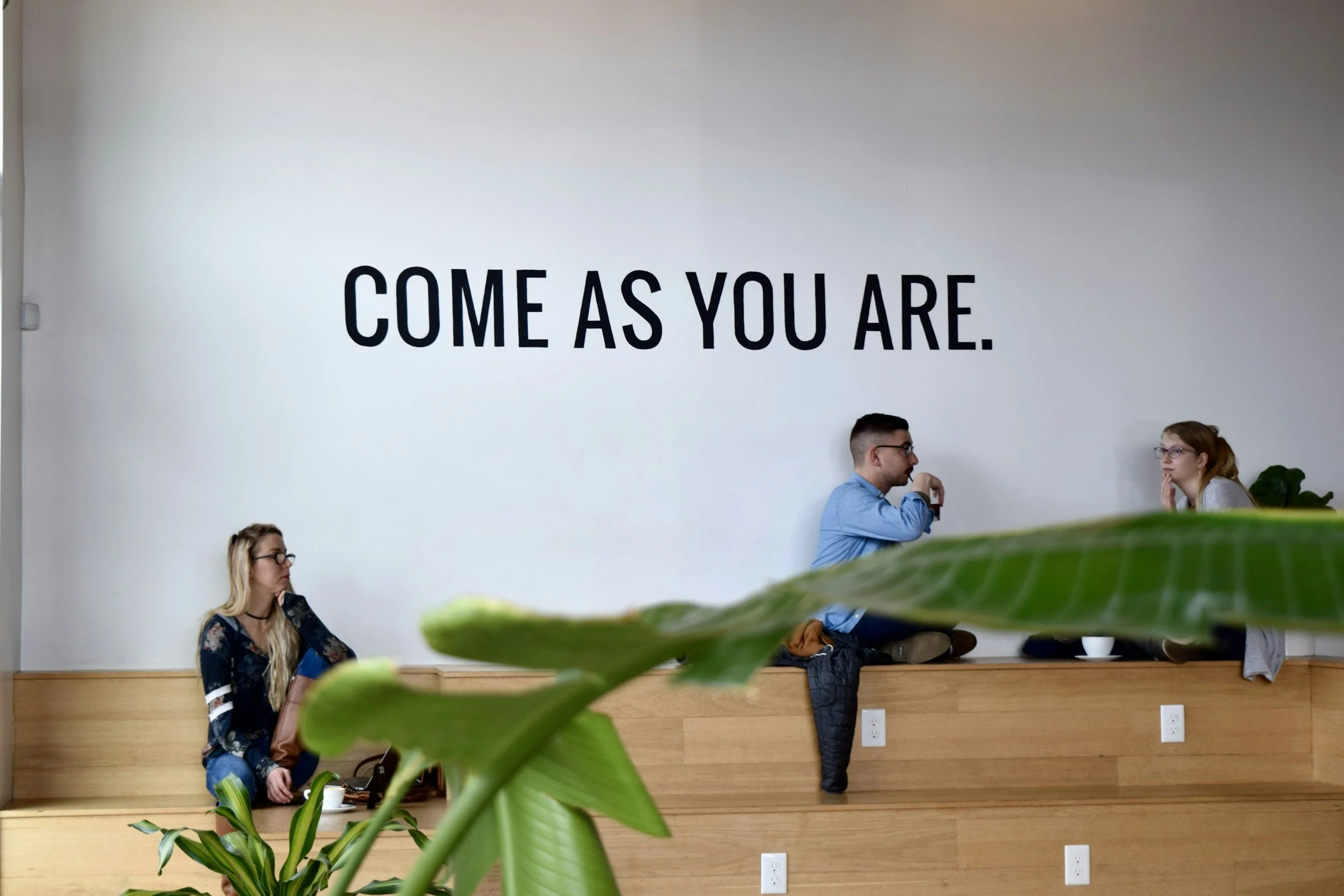 people sitting together in a simple room with big plant. the quote says "come as you are"