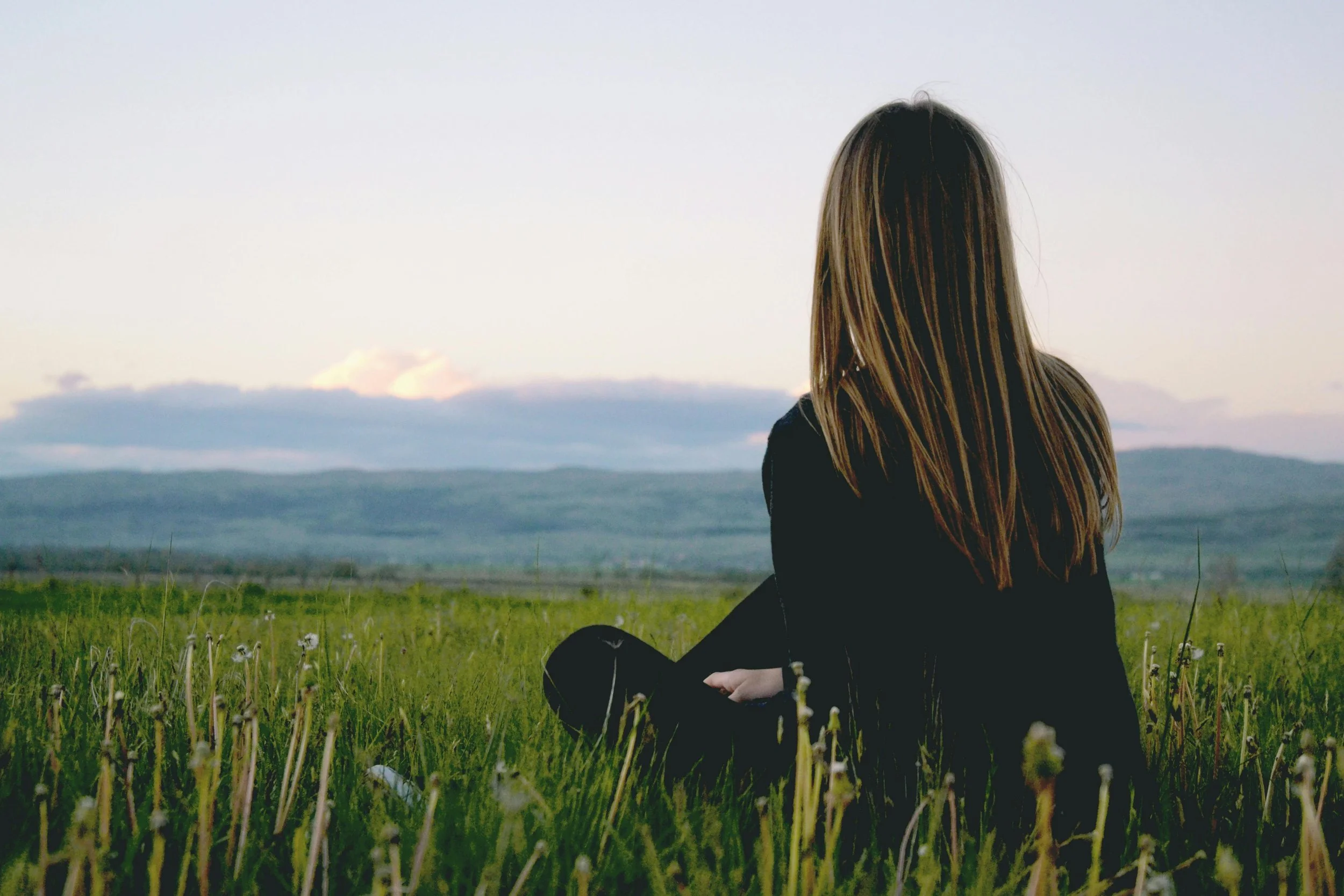 A woman with long hair sitting in a grassy field, facing away, with mountains and a cloudy sky in the background.