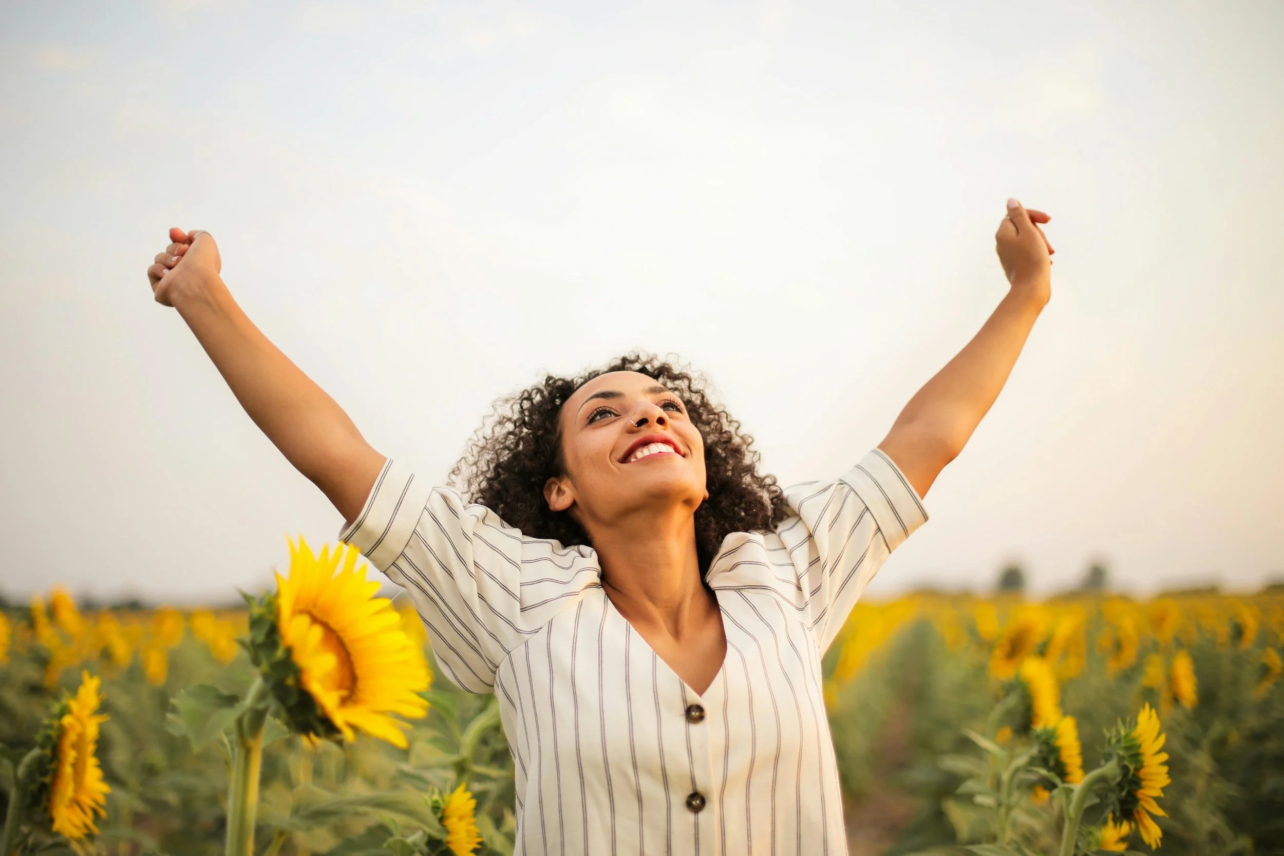 A woman with curly hair smiling and raising her arms in a sunflower field on a cloudy day.