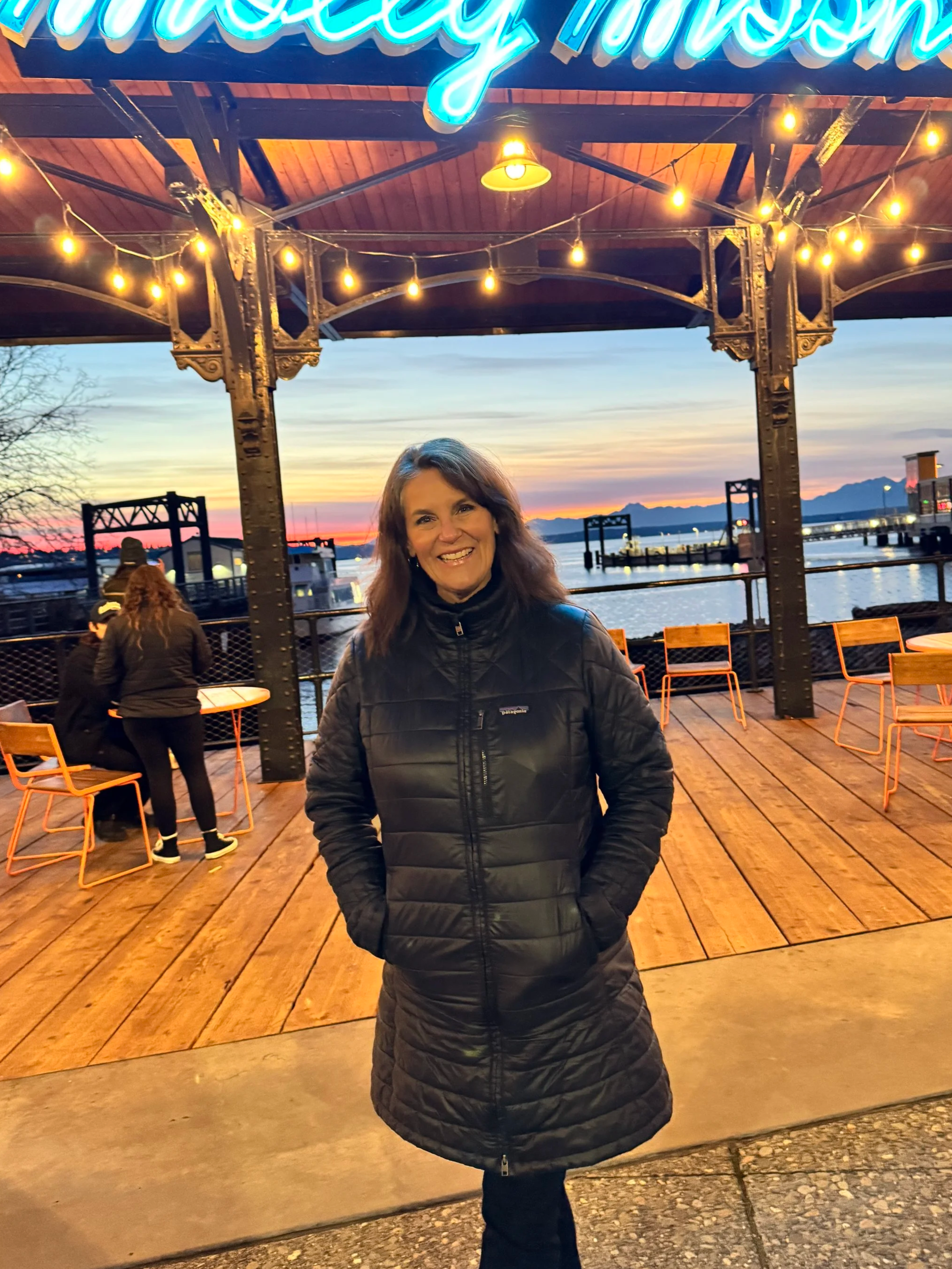 A woman wearing a black quilted jacket smiling at the camera on a wooden outdoor deck with string lights hanging overhead, with a sunset over a body of water and mountains in the background.