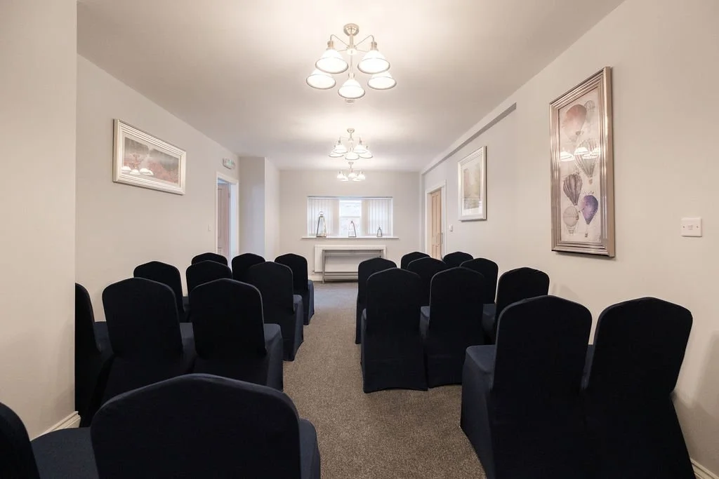A small conference or meeting room with rows of black chairs facing a windowed wall with a white fireplace mantel, decorated with framed artwork on the beige walls and ceiling lights.