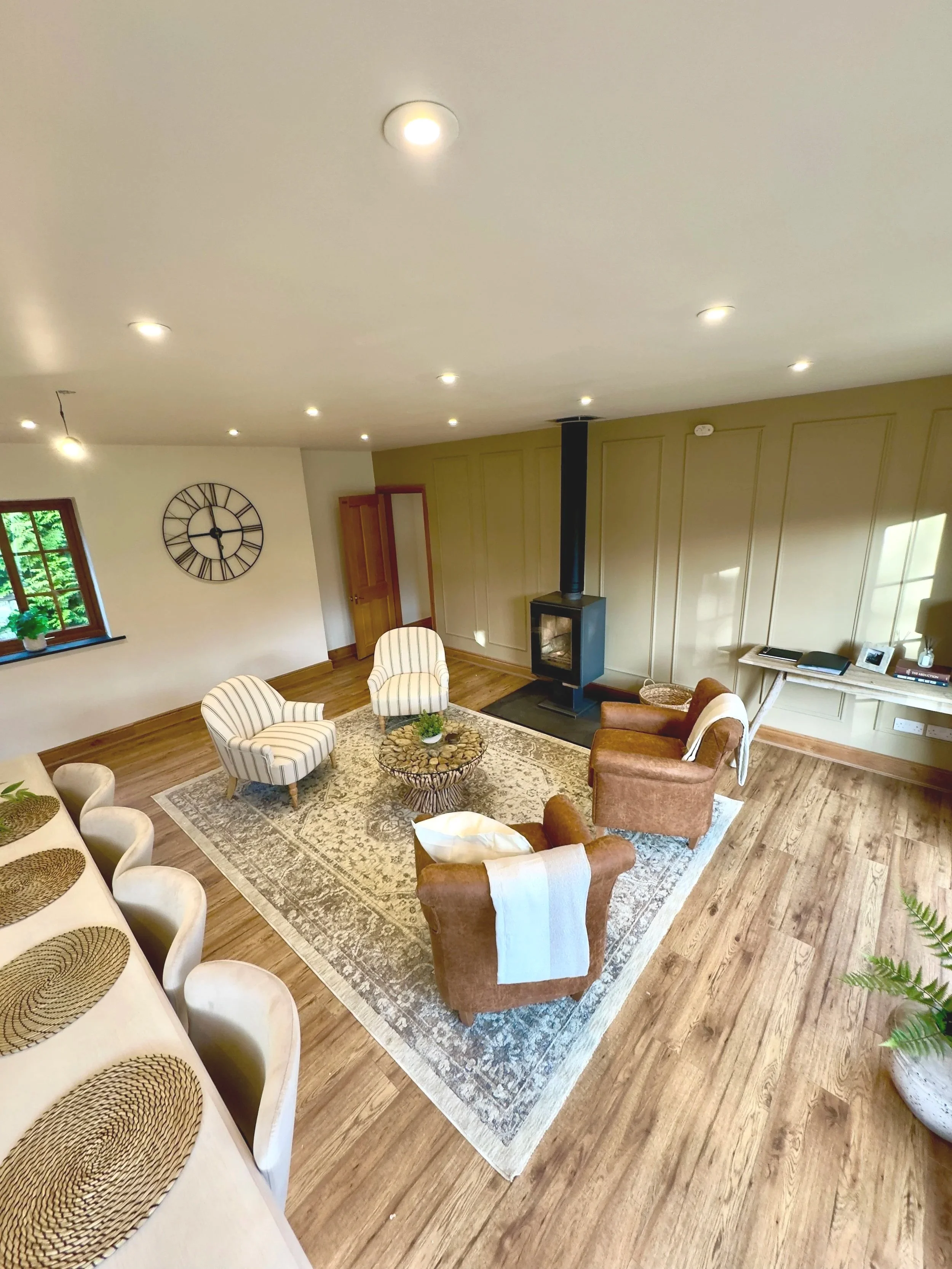 A cosy and inviting living room featuring a striking black fireplace set against a panelled accent wall. Oak flooring provides a warm foundation, layered with a large patterned rug that adds texture and visual interest. Seating includes two sleek str