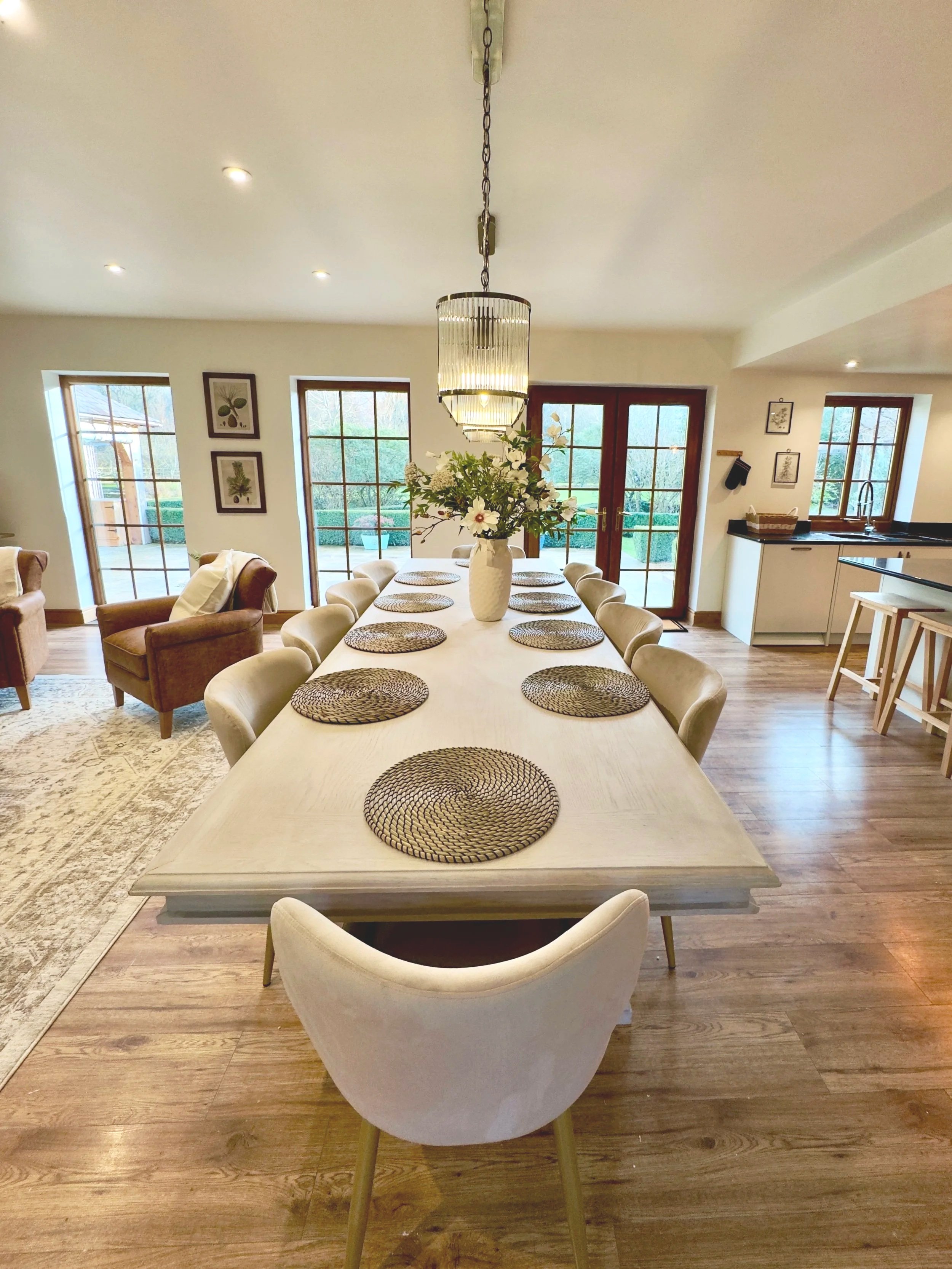 A bright and airy dining space featuring a large white table surrounded by ten comfortable chairs. Woven placemats add natural texture, while a vase of fresh flowers sits at the centre, bringing life and colour to the table. A pendant light hangs ove