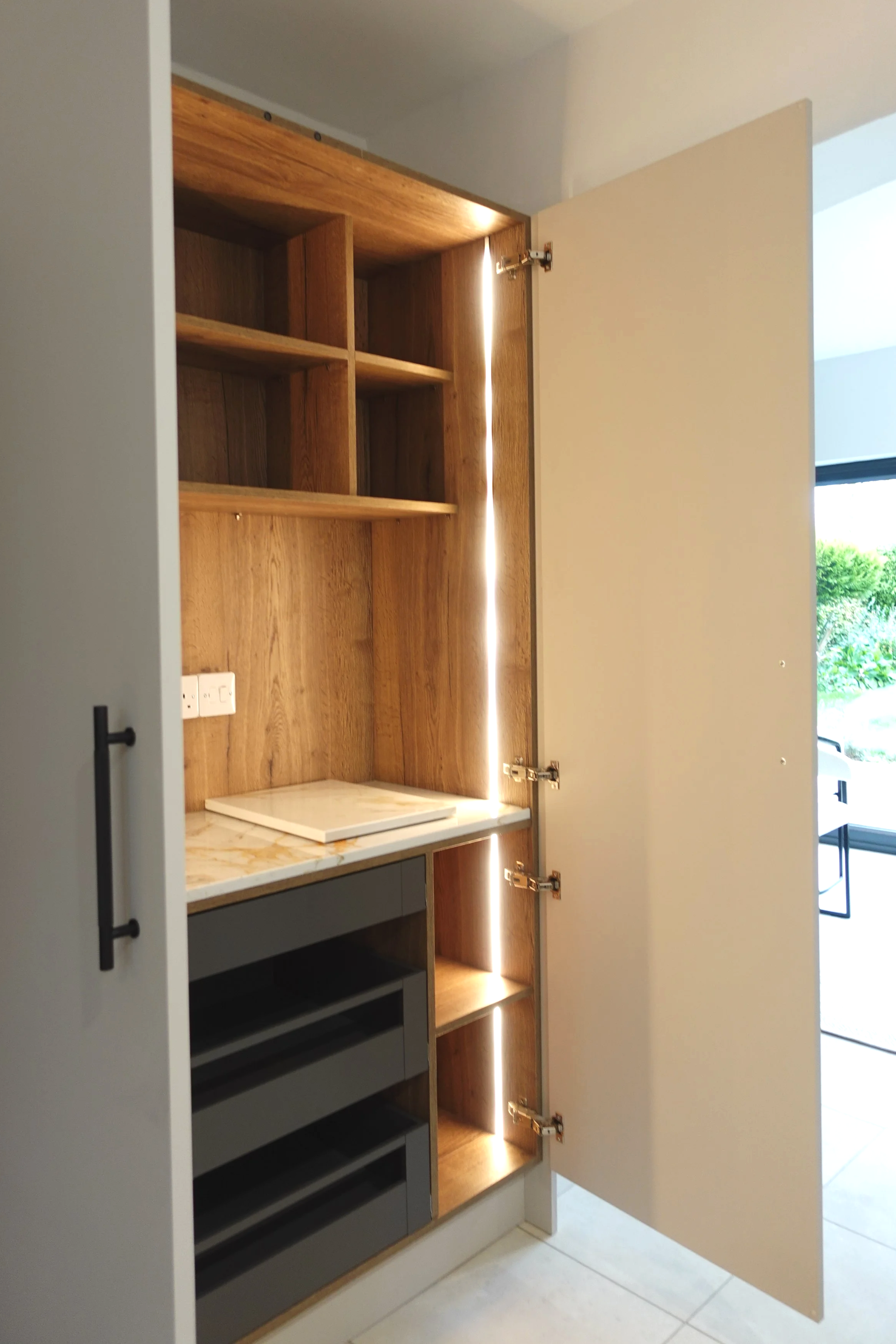 A beautifully crafted built-in pantry cabinet designed to combine practicality with refined detail. The tall unit opens to reveal warm oak interior shelving with integrated LED lighting, creating a soft glow that highlights the natural wood grain. Mu