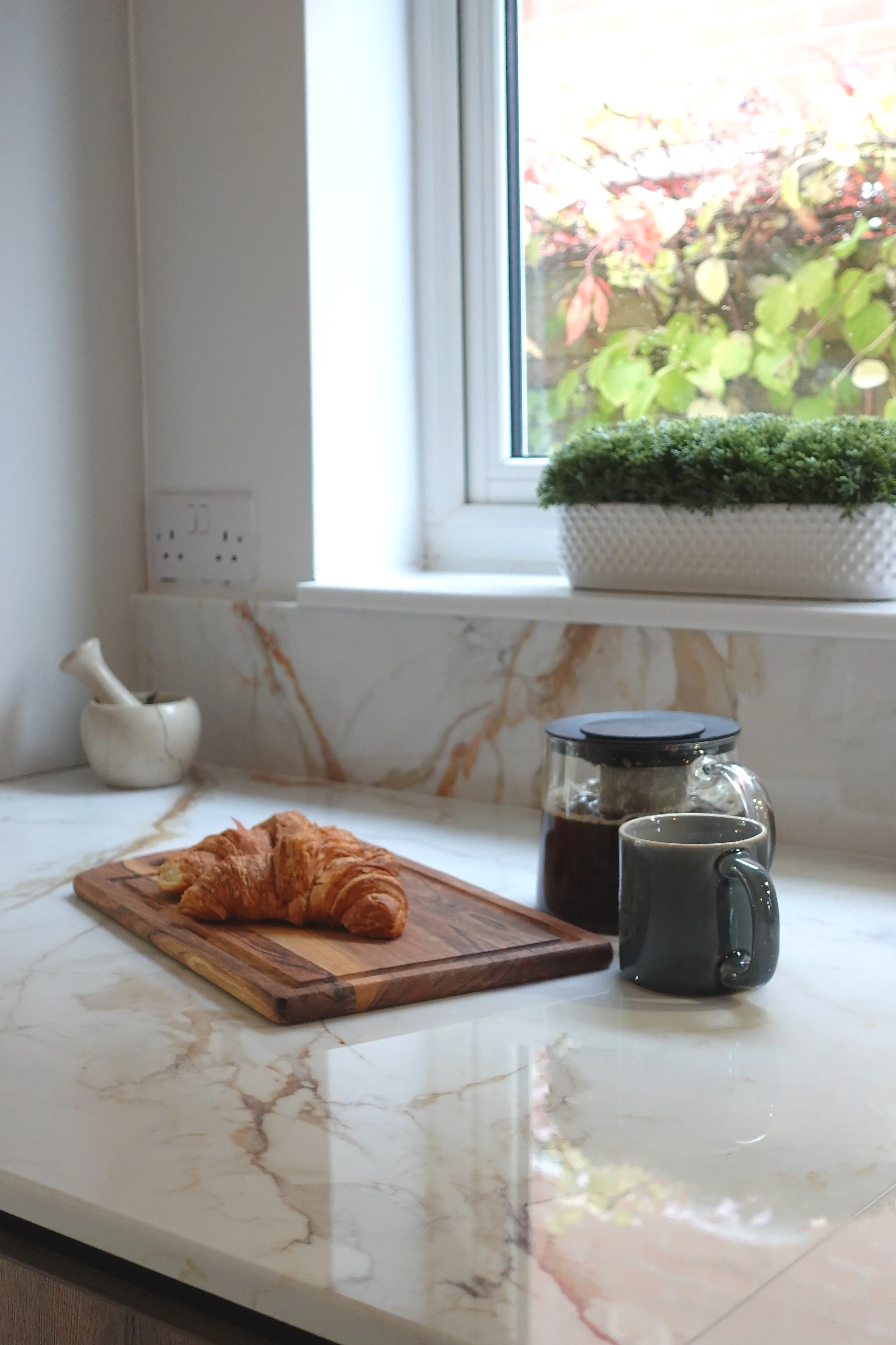 A bright and inviting kitchen worktop styled with simple, elegant details. The marble-effect surface with warm natural veining reflects the daylight from the nearby window, creating a fresh and airy feel. A wooden board with freshly baked croissants 