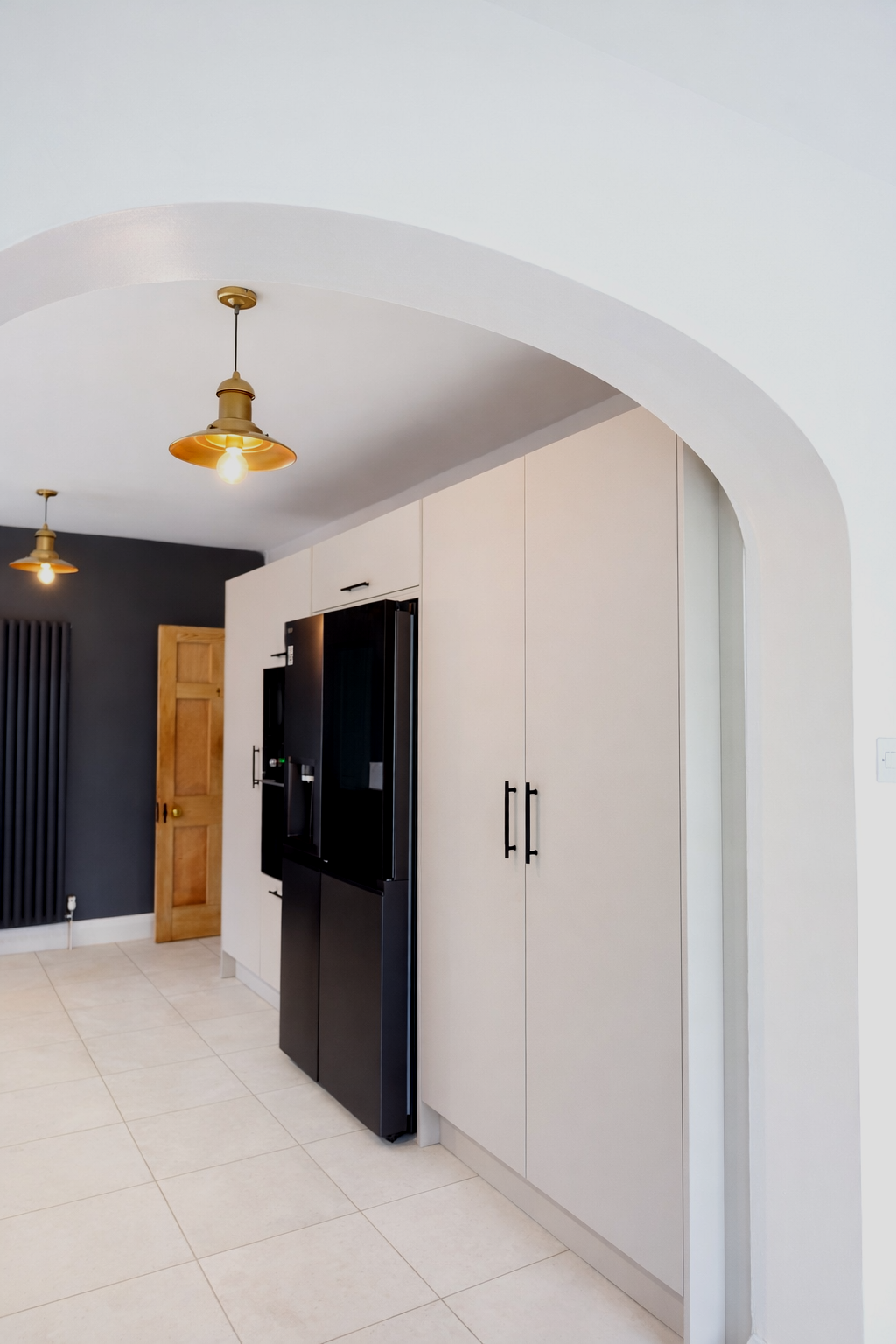 A clean and elegant kitchen corridor framed by a soft architectural arch, creating a welcoming transition into the space. The design features tall, minimalist cabinetry in a light finish paired with sleek black handles and integrated appliances for a