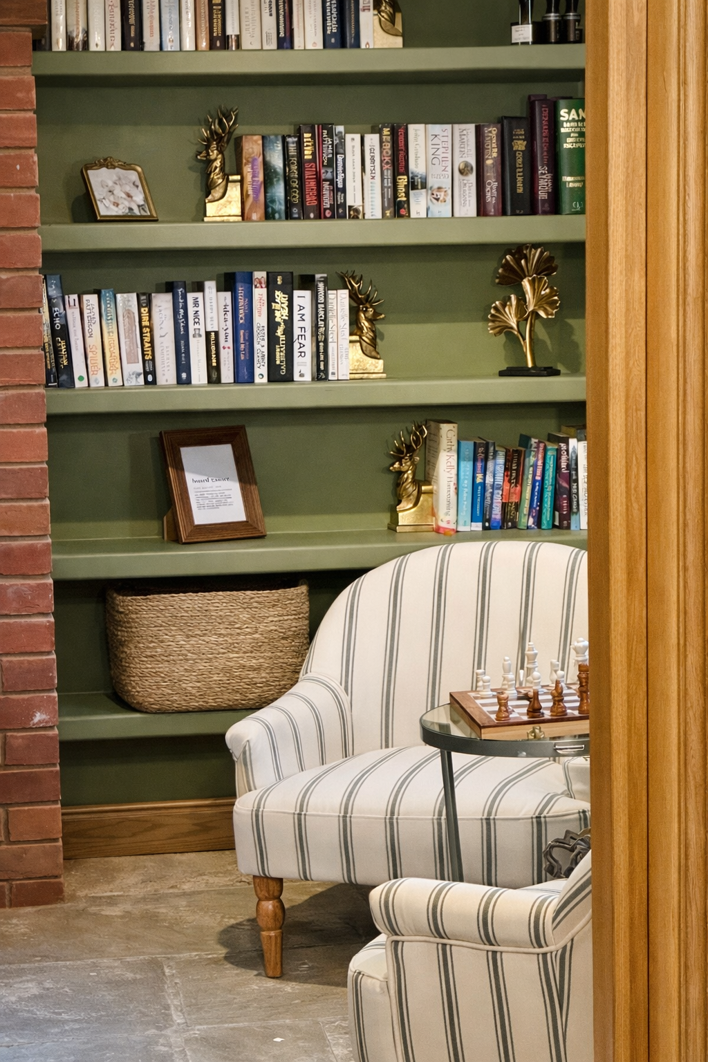 A cosy, characterful reading nook featuring bespoke olive green built-in shelving, styled with curated books, brass accents, and decorative objects. Set against exposed brick detailing, the space is softened by a classic striped armchair and warm tim