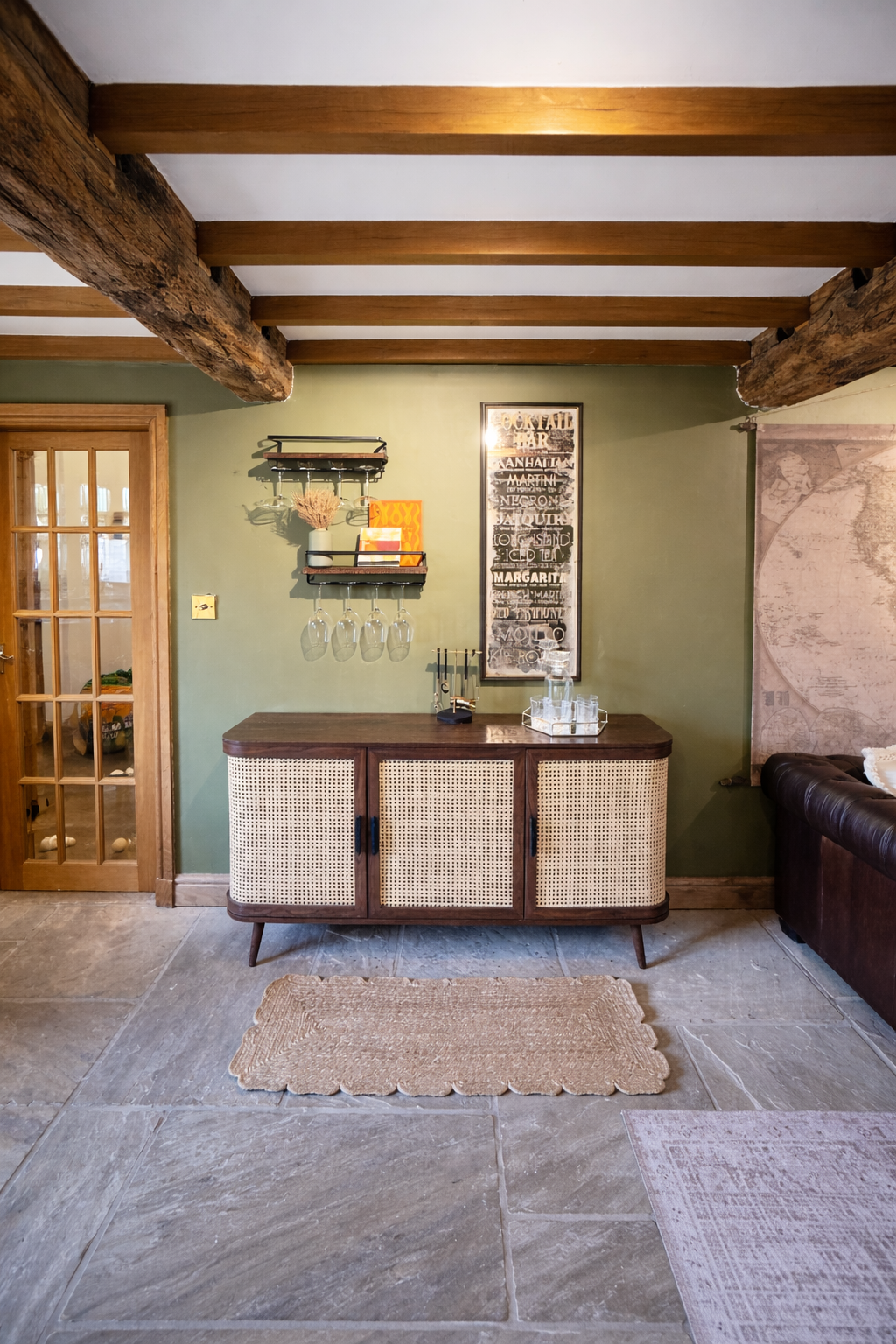 A warm, characterful entertaining space defined by exposed timber ceiling beams and a rich olive green backdrop. A mid-century inspired sideboard with woven cane fronts anchors the room, paired with bespoke wall-mounted shelving that neatly displays 