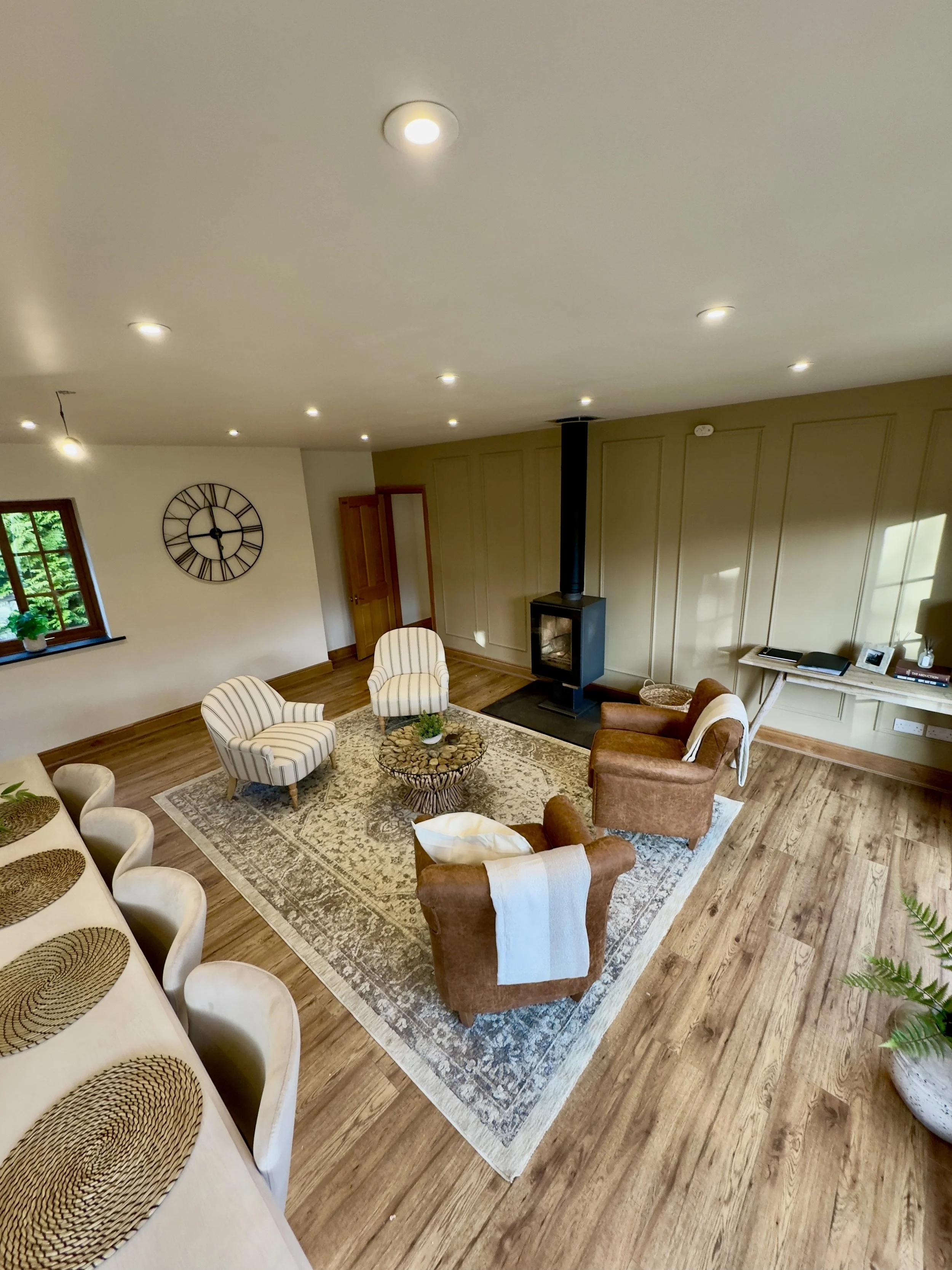 A cosy and inviting living room featuring a striking black fireplace set against a panelled accent wall. Oak flooring provides a warm foundation, layered with a large patterned rug that adds texture and visual interest. Seating includes two sleek str